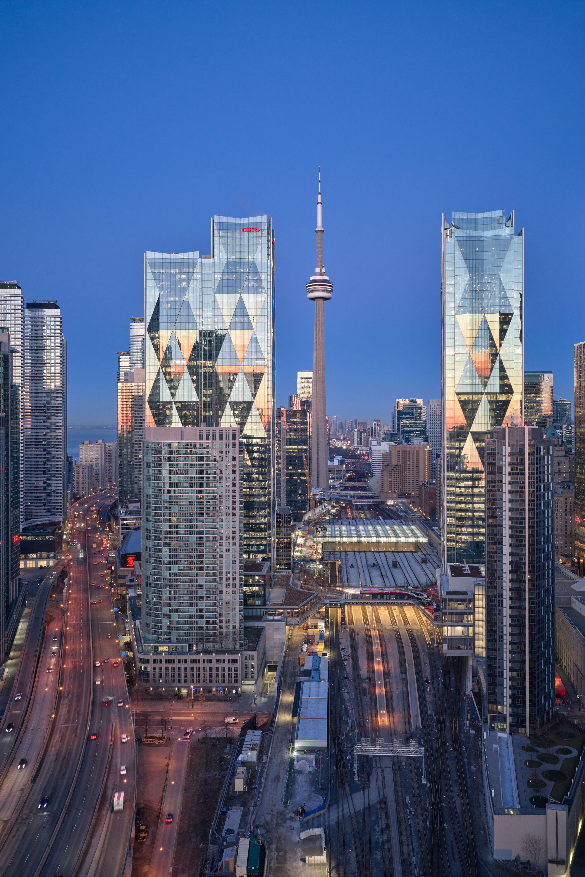 Twilight view of twin towers with diamond-patterned facades flanking the rail corridor and transmission tower
