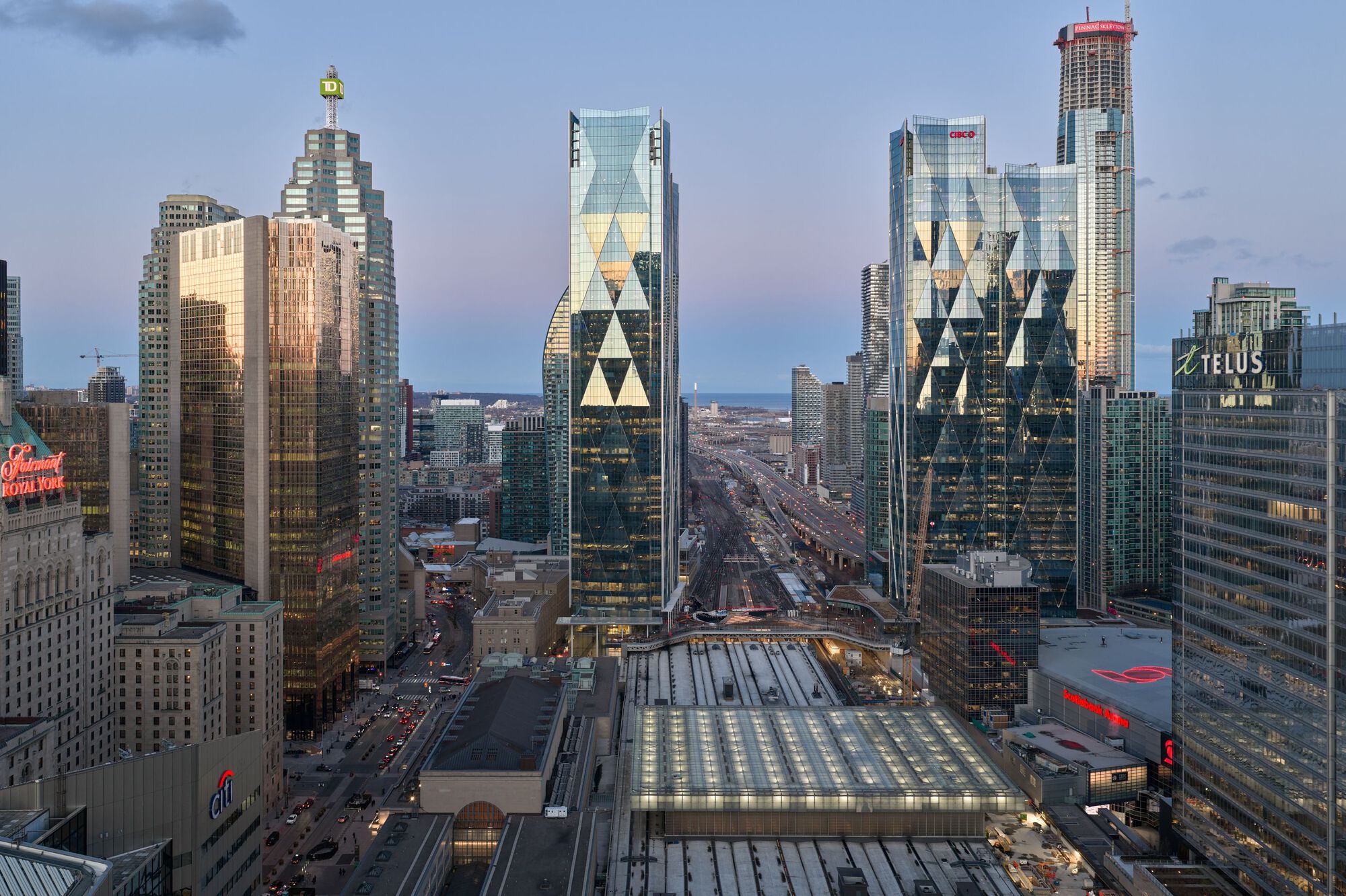 Elevated dusk view of the towers and rail shed with illuminated glass roofs and surrounding cityscape