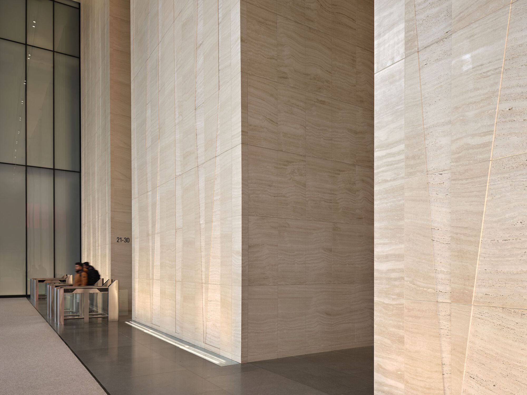 Corner of a backlit travertine wall panel with a person seated at security turnstiles