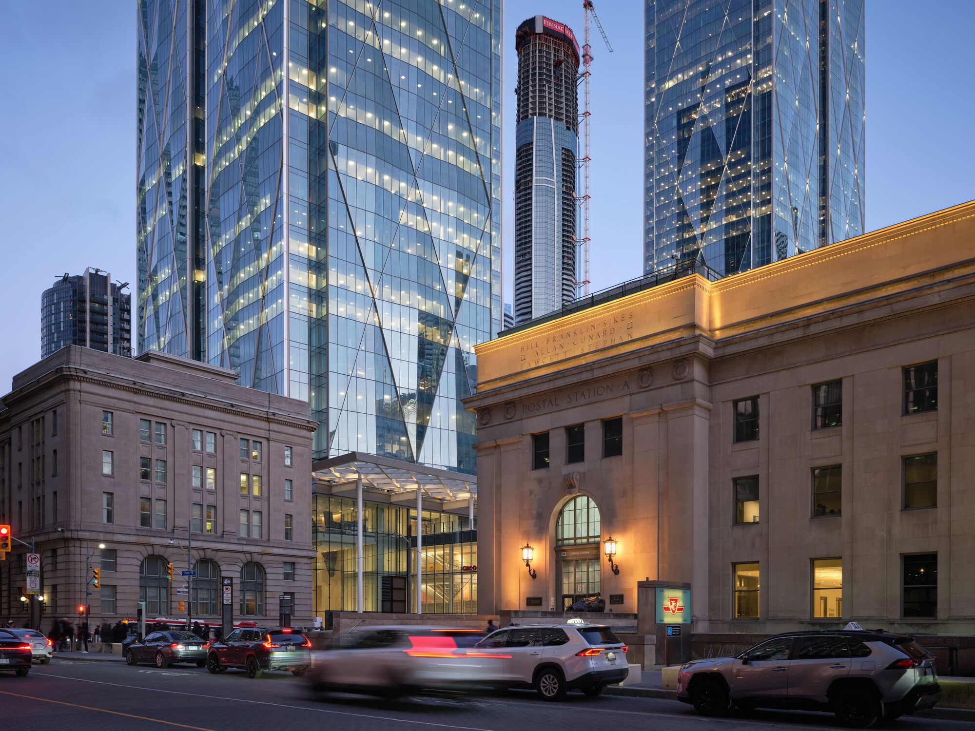 Glass tower base meeting a heritage arched facade at dusk with vehicle light trails