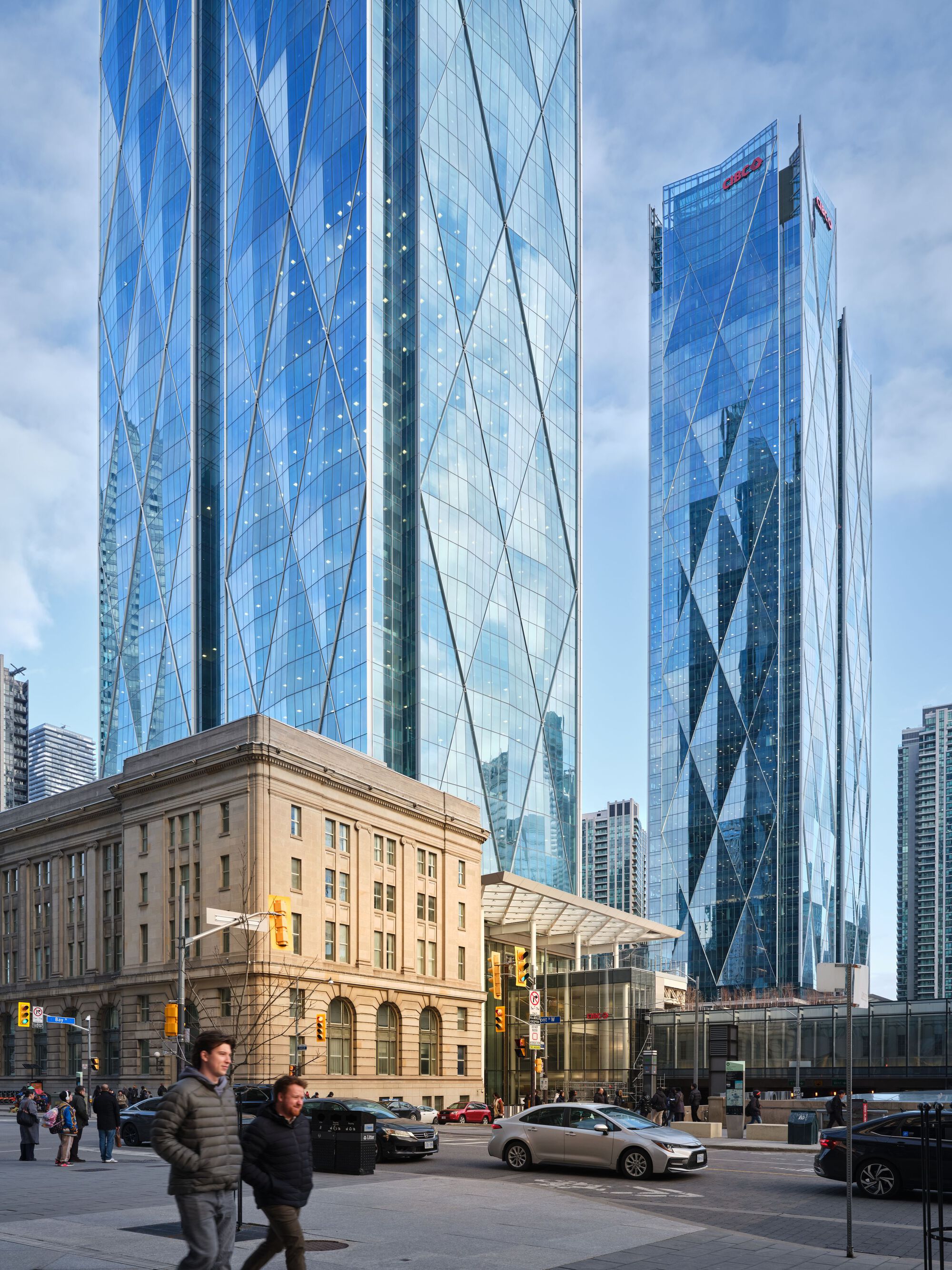 Street-level view of the glass towers behind a limestone heritage building with pedestrians crossing the intersection