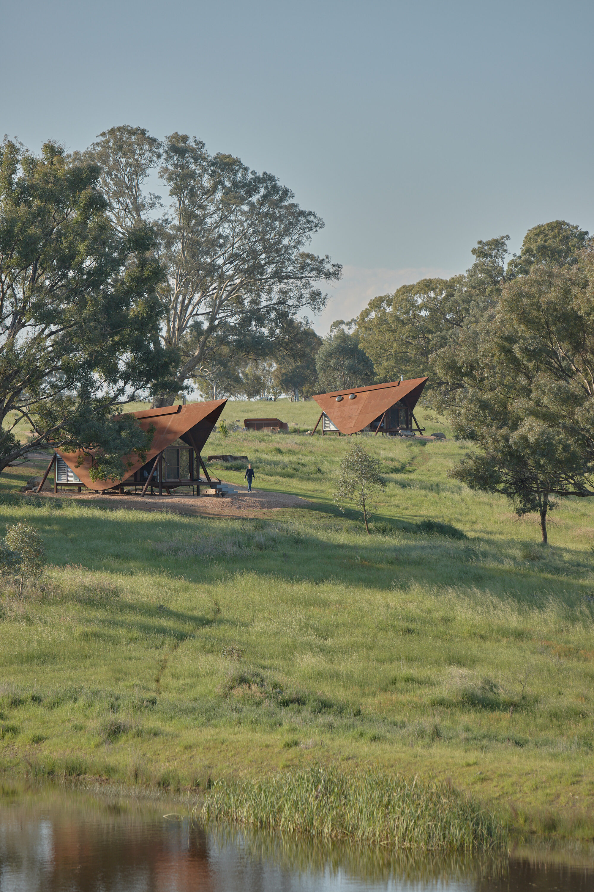 Three tented cabins with rust-colored roofs scattered across a sloping grassy field with a pond
