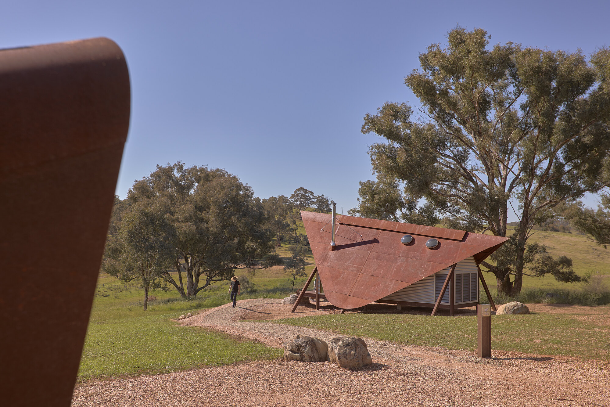 Weathered steel canopy sheltering a glazed cabin wall with a figure walking on gravel beneath