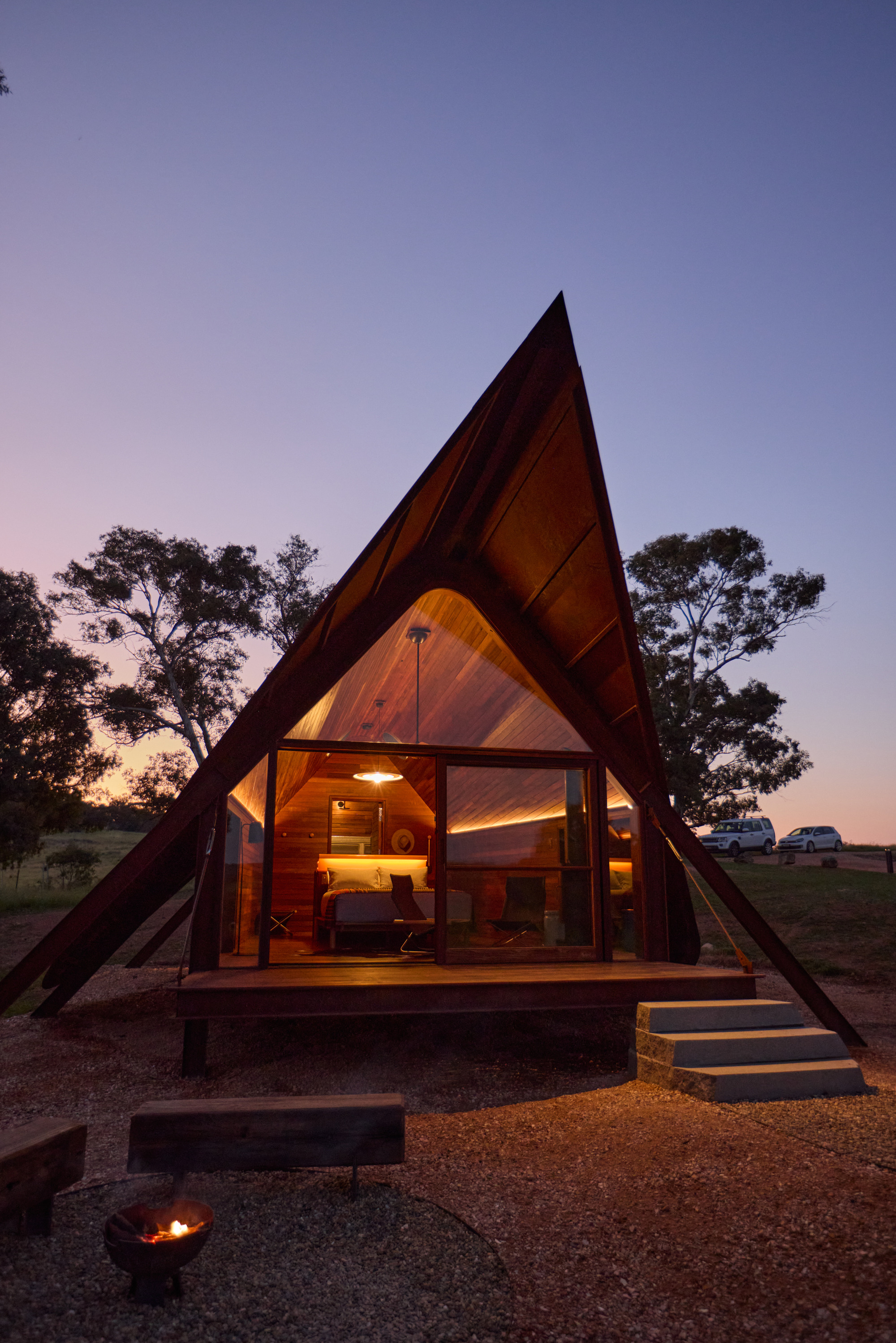 Steeply pitched timber frame cabin with glazed gable end illuminated at dusk beside a fire pit