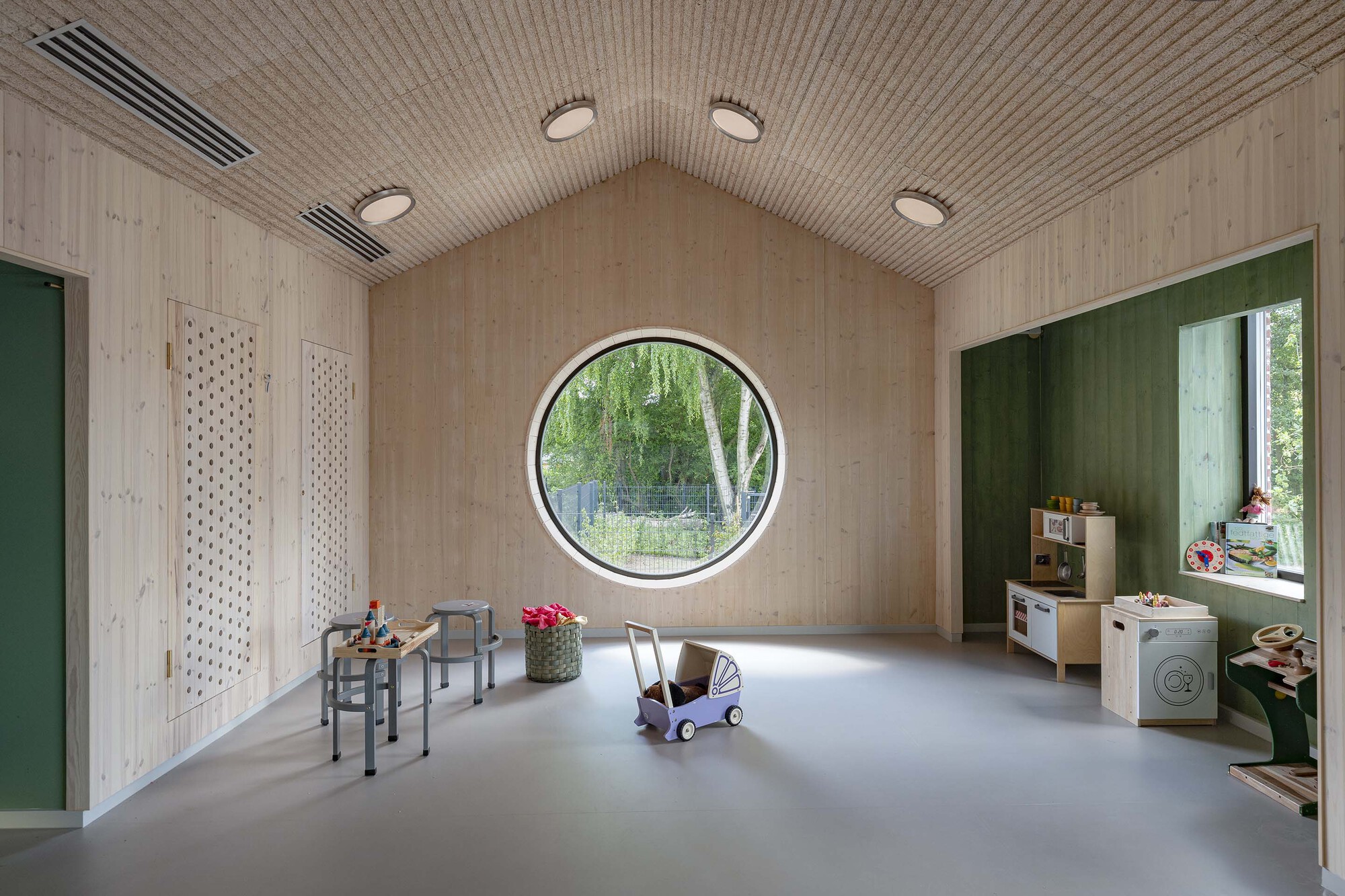 Playroom with pale timber walls, ribbed ceiling and circular window framing birch trees outside