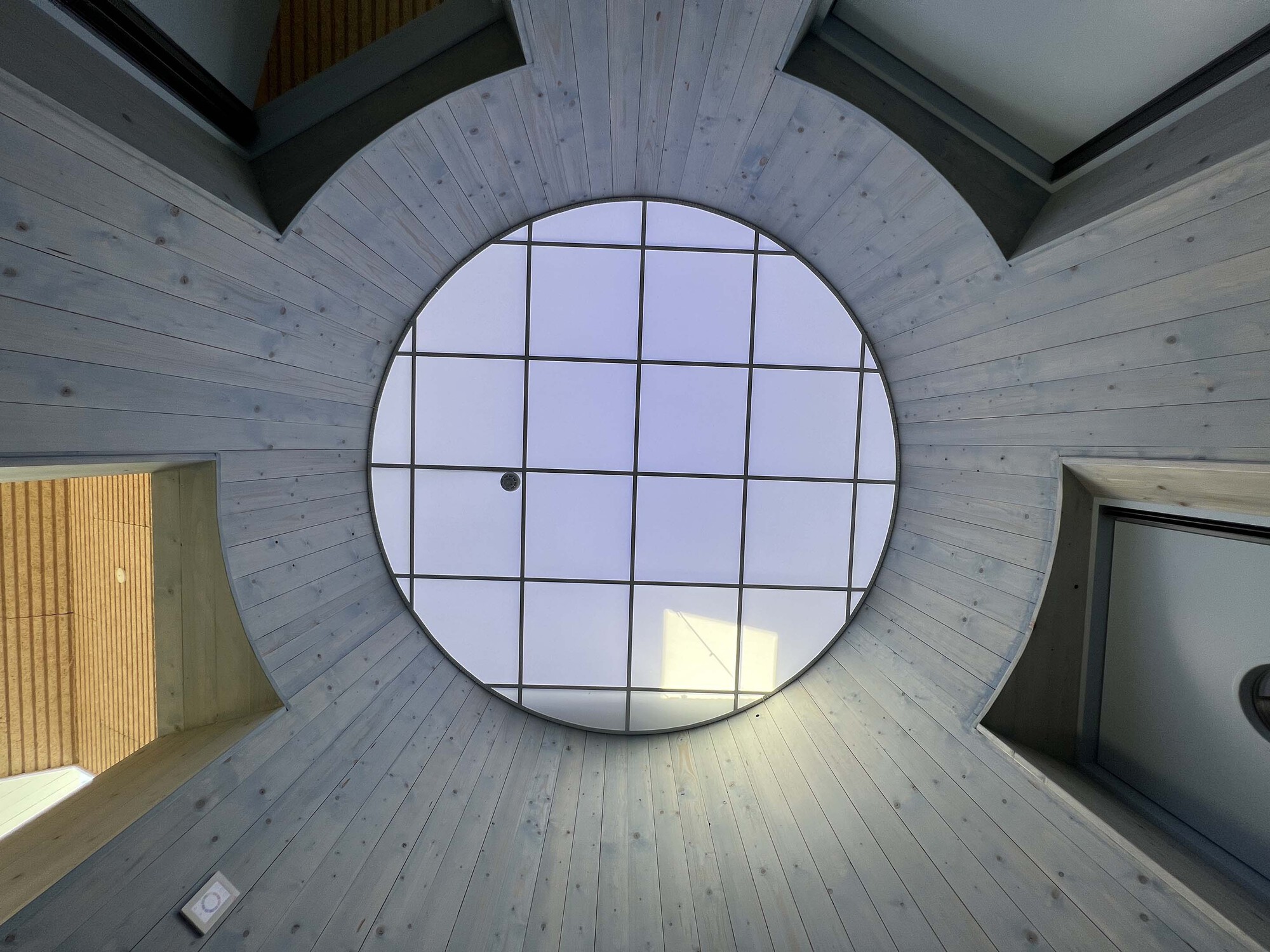 Upward view of a circular gridded skylight framed by whitewashed timber planks under an overcast sky