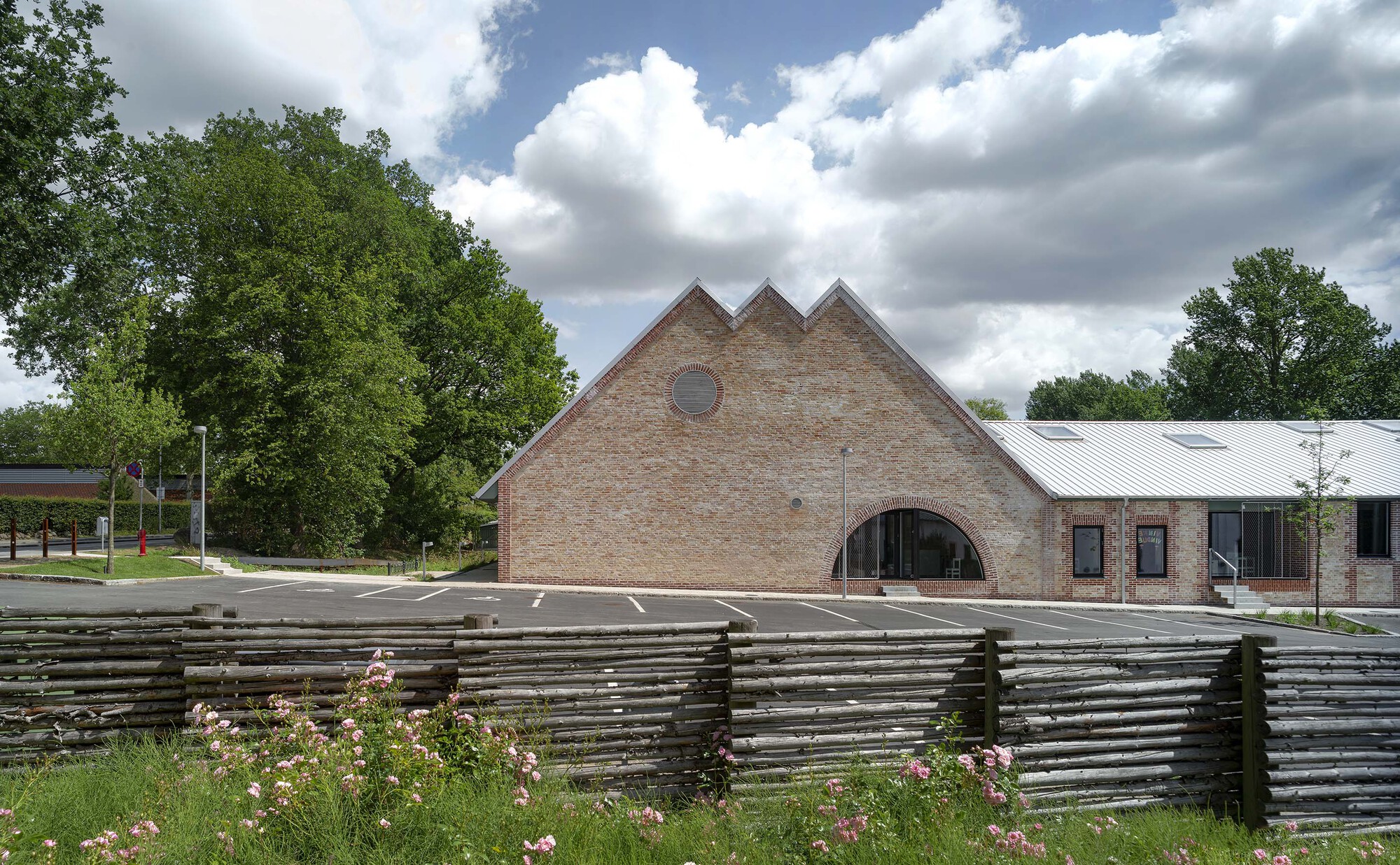 Triple-gabled brick facade with arched entrance behind a low woven timber fence and planted bed