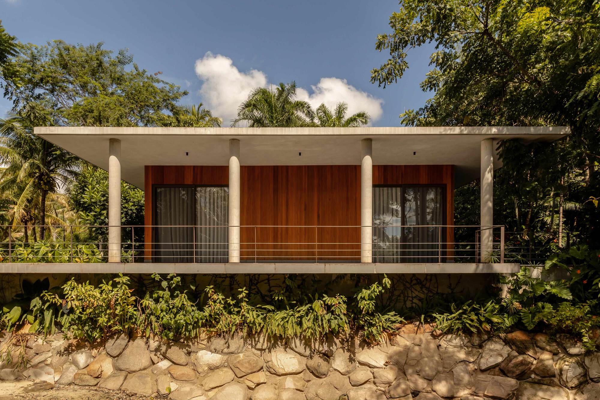 Elevated concrete volume with timber-clad facade and cable railing above a stone-retained embankment surrounded by palms