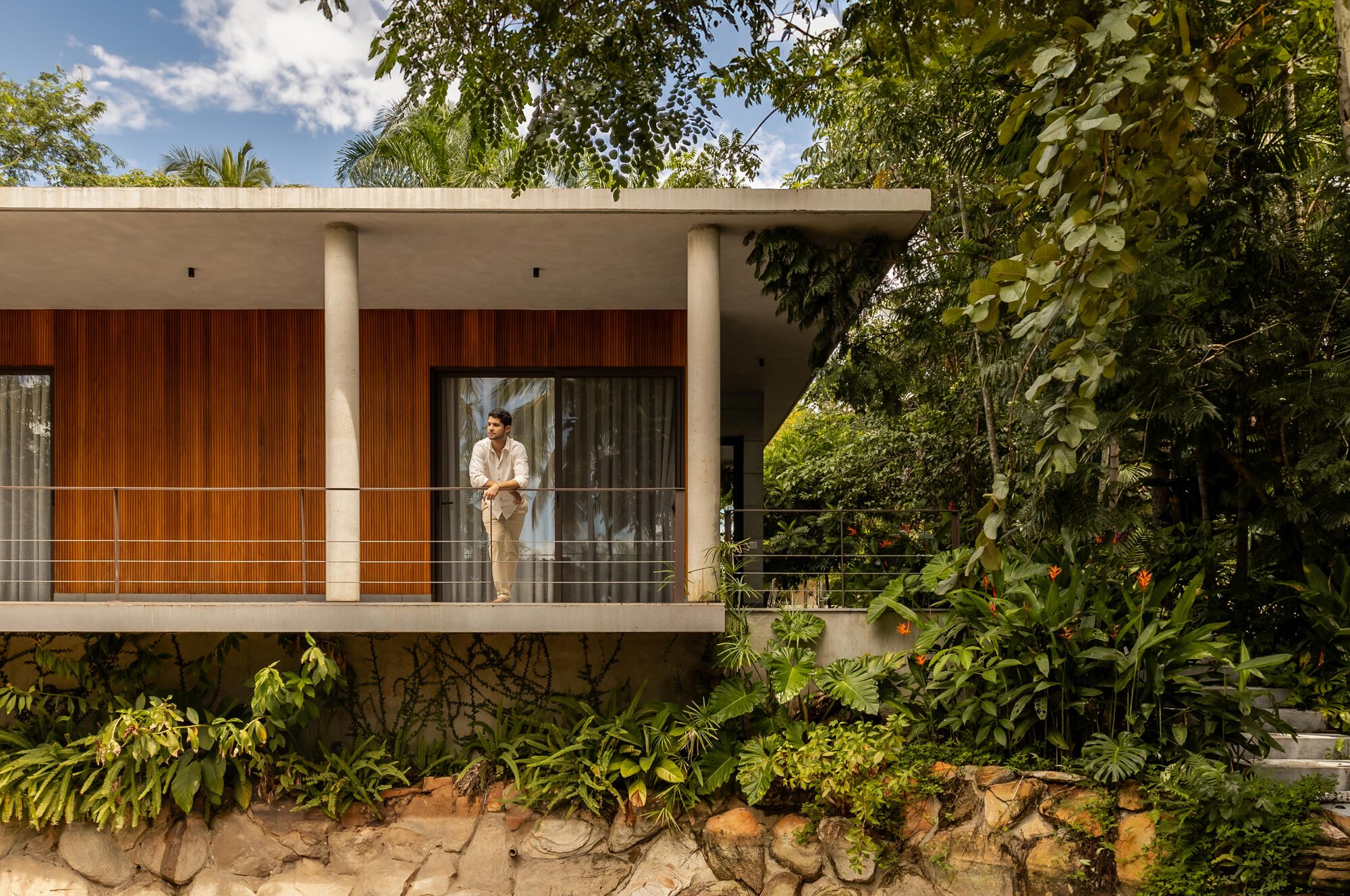 Cable-railed terrace with person standing behind vertical timber panels amid dense tropical vegetation