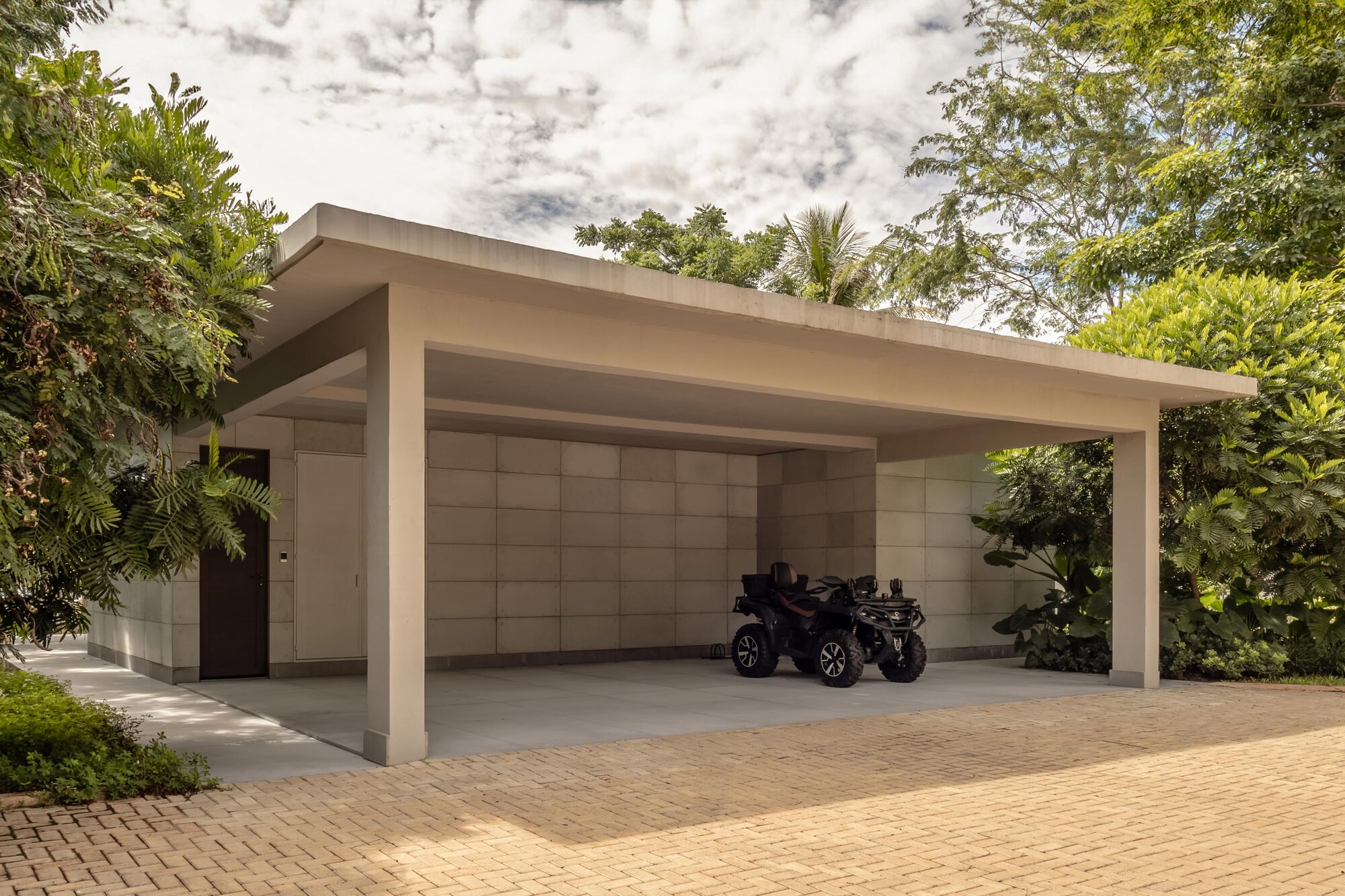 Open carport with flat concrete roof, paneled wall and brick paving surrounded by tropical vegetation