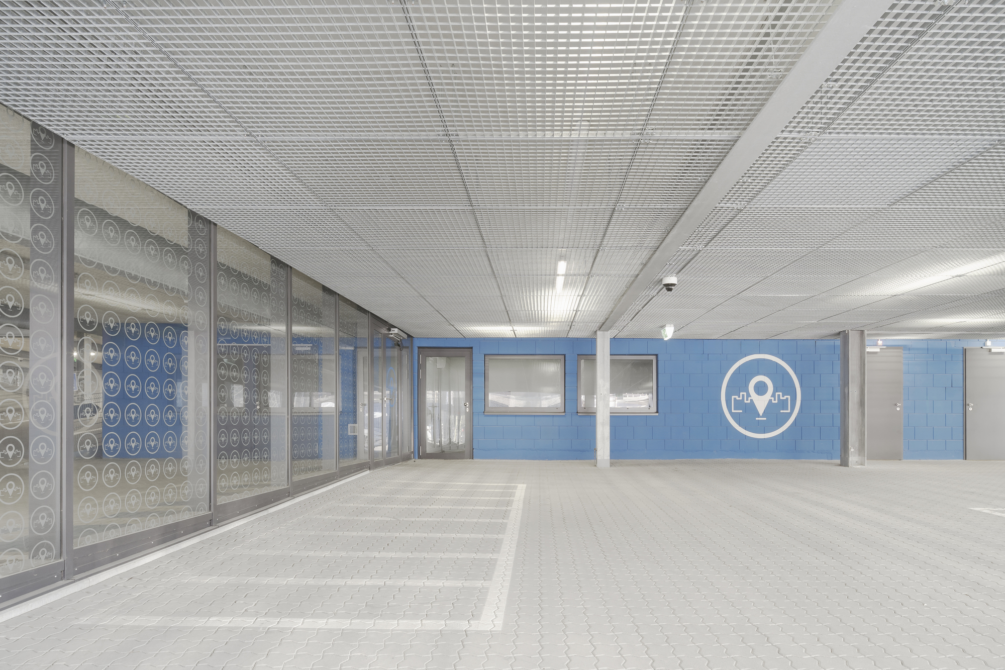 Interior parking level with perforated glass panels and blue painted wall beneath a metal mesh ceiling