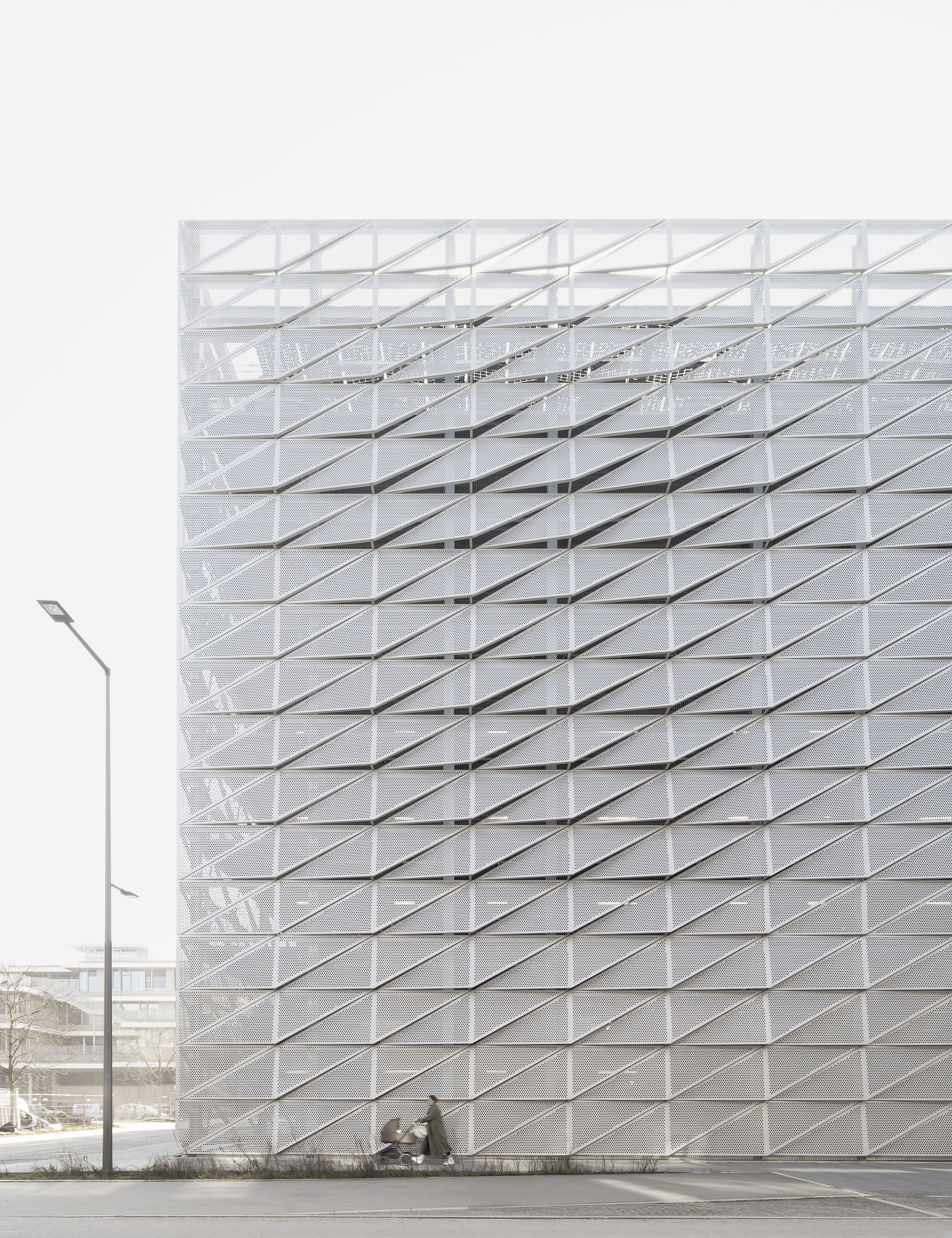 White folded panel facade with diagonal grid pattern and pedestrians at ground level under overcast sky