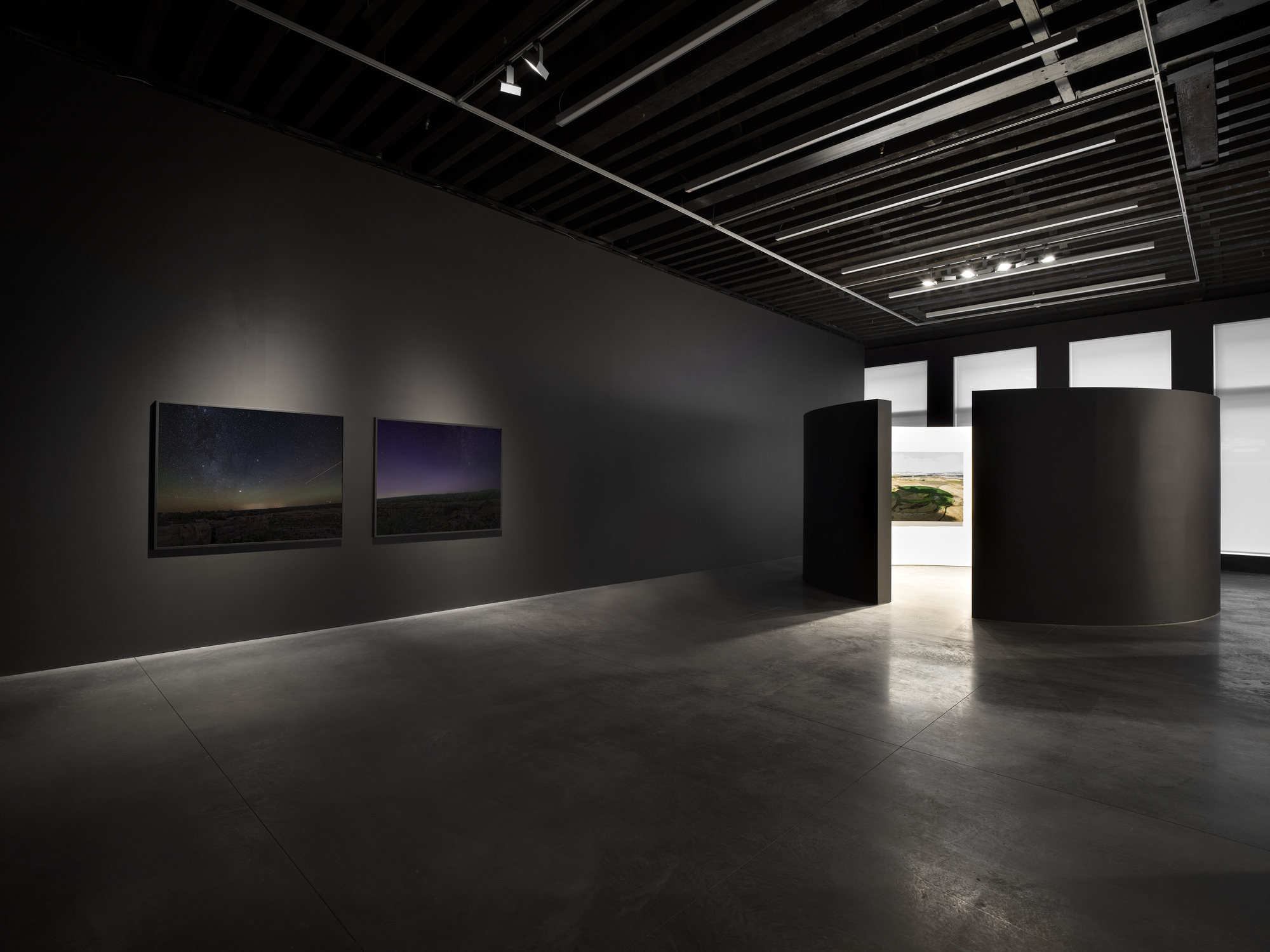 Dark gallery space with charcoal walls, exposed black ceiling joists and cylindrical partition element