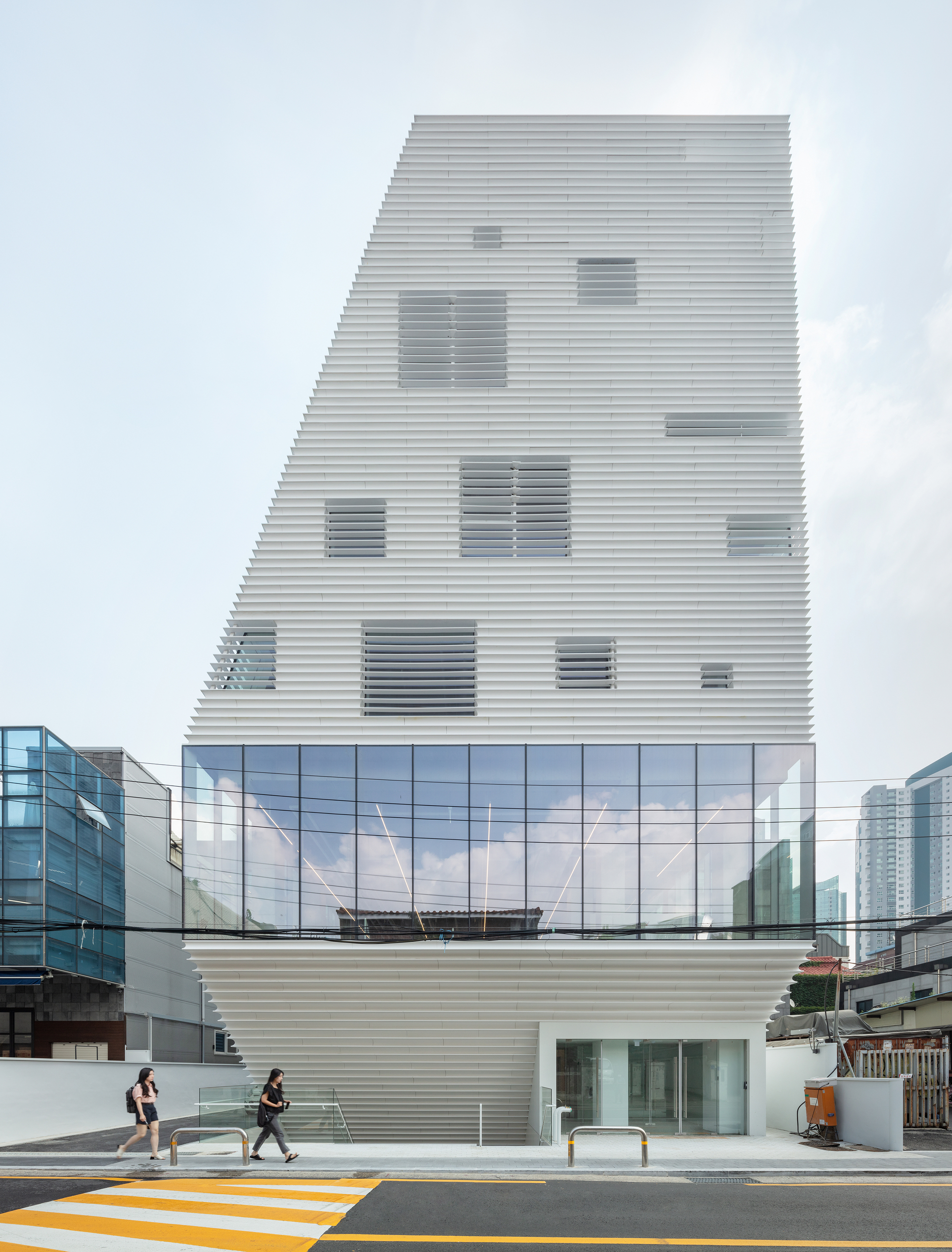 Street view of the white horizontal louver facade with irregular window openings and pedestrians crossing below