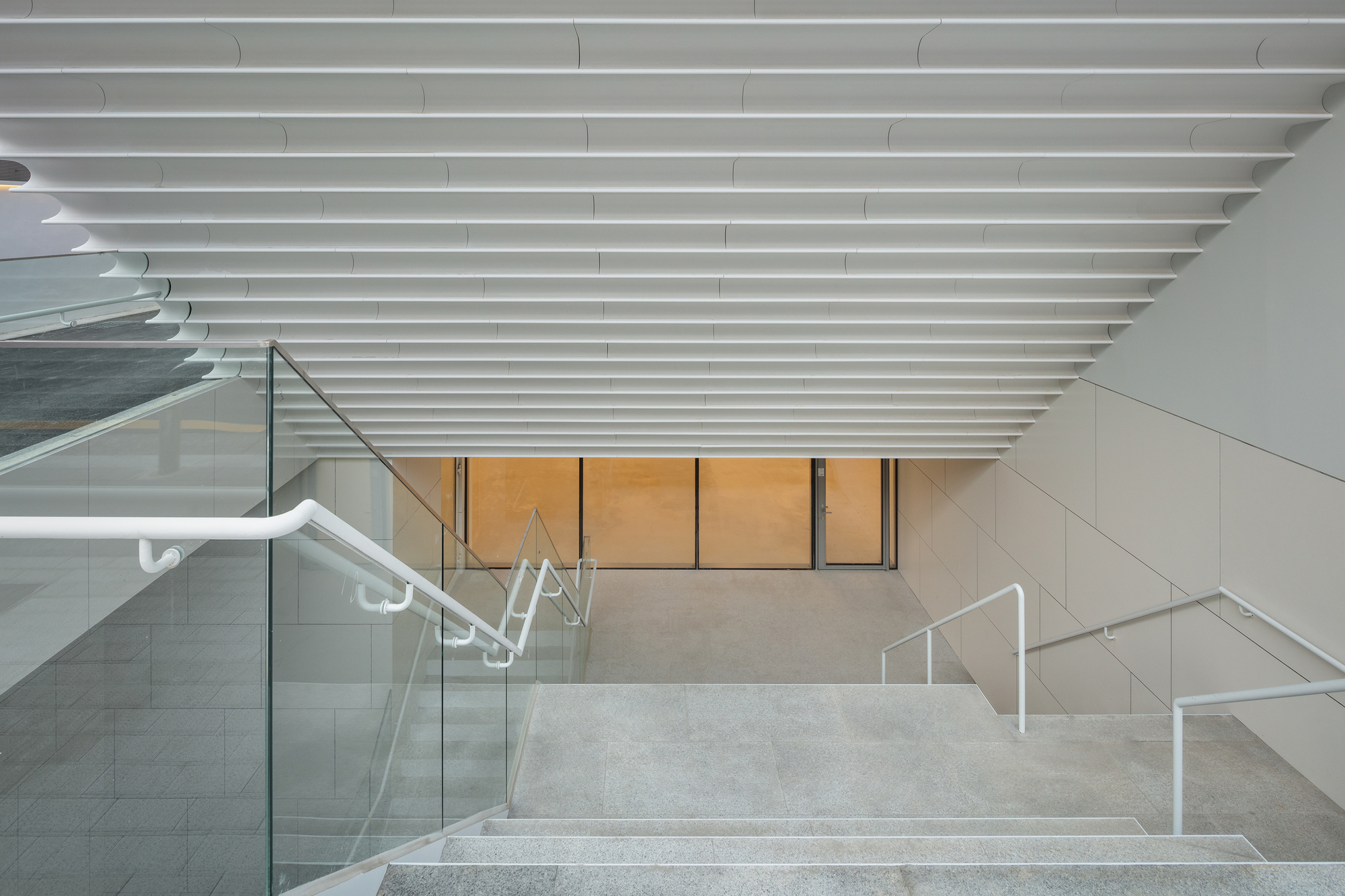 Interior staircase with glass balustrade and white railings descending beneath horizontal slatted ceiling