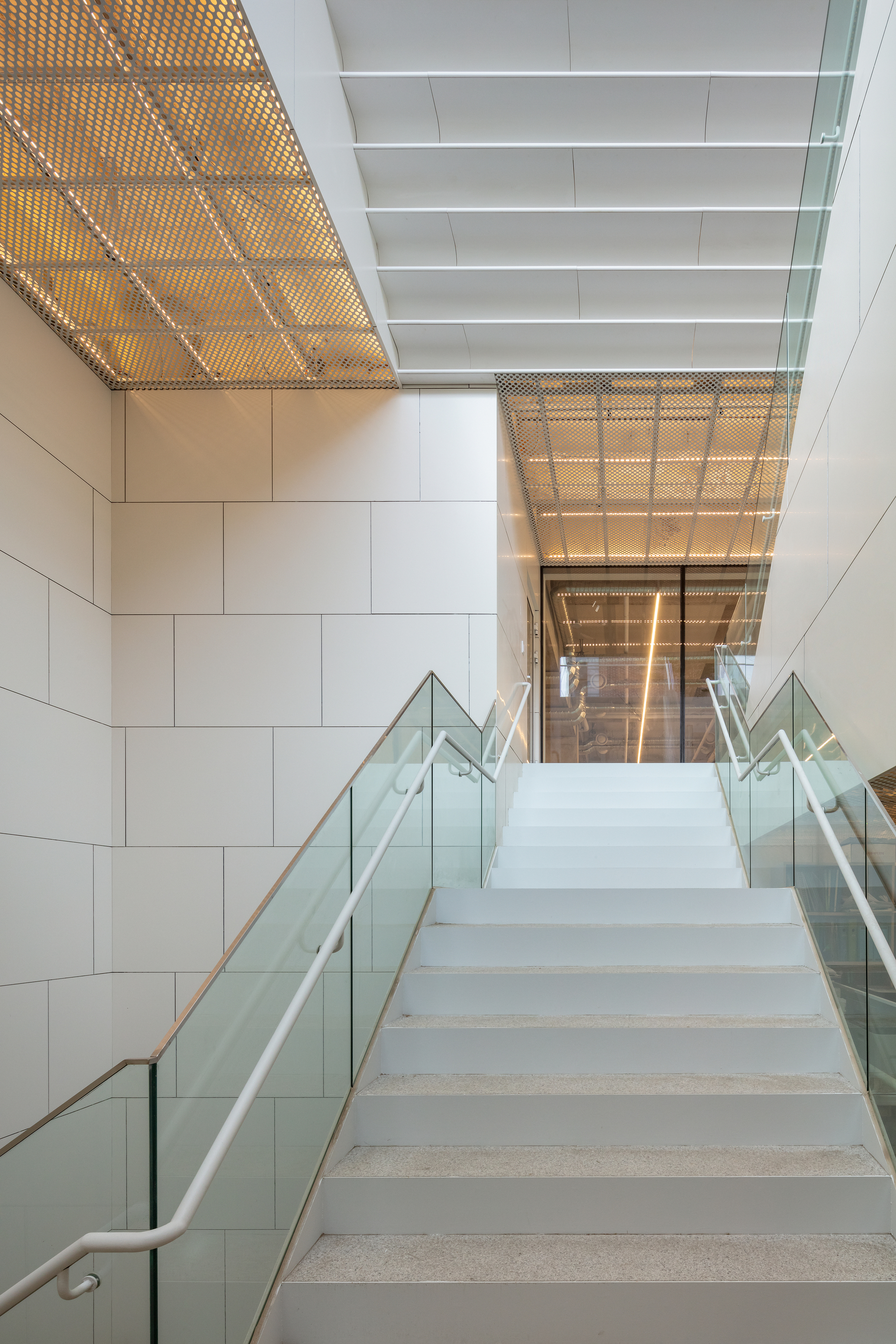 Stairwell with perforated metal ceiling panels and glass balustrades lit by warm afternoon light