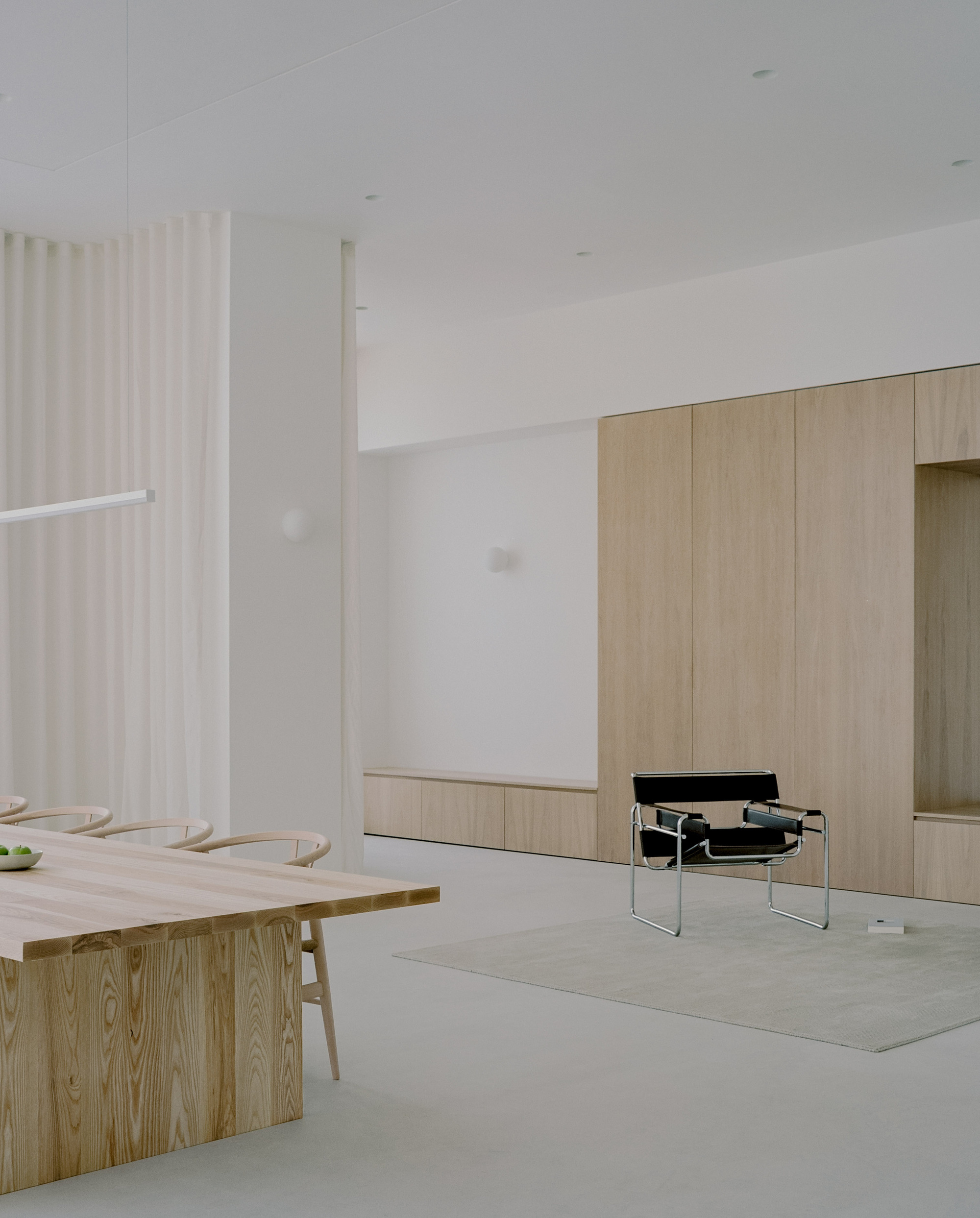 Open living space with timber dining table and built-in cabinetry beneath white ceiling and sheer curtains