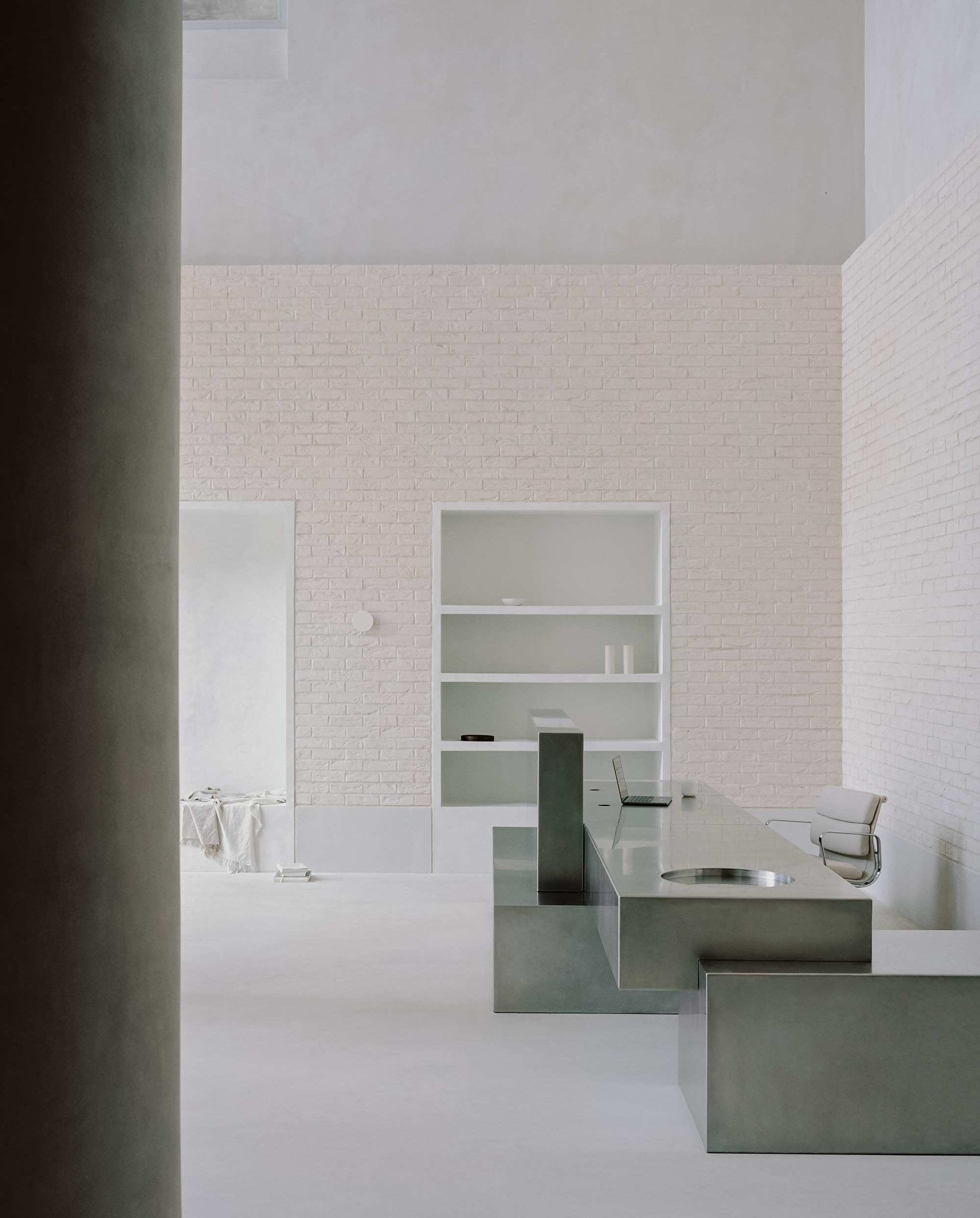 Minimalist workspace with white painted brick walls, built-in shelving, and a stainless steel desk under soft natural light