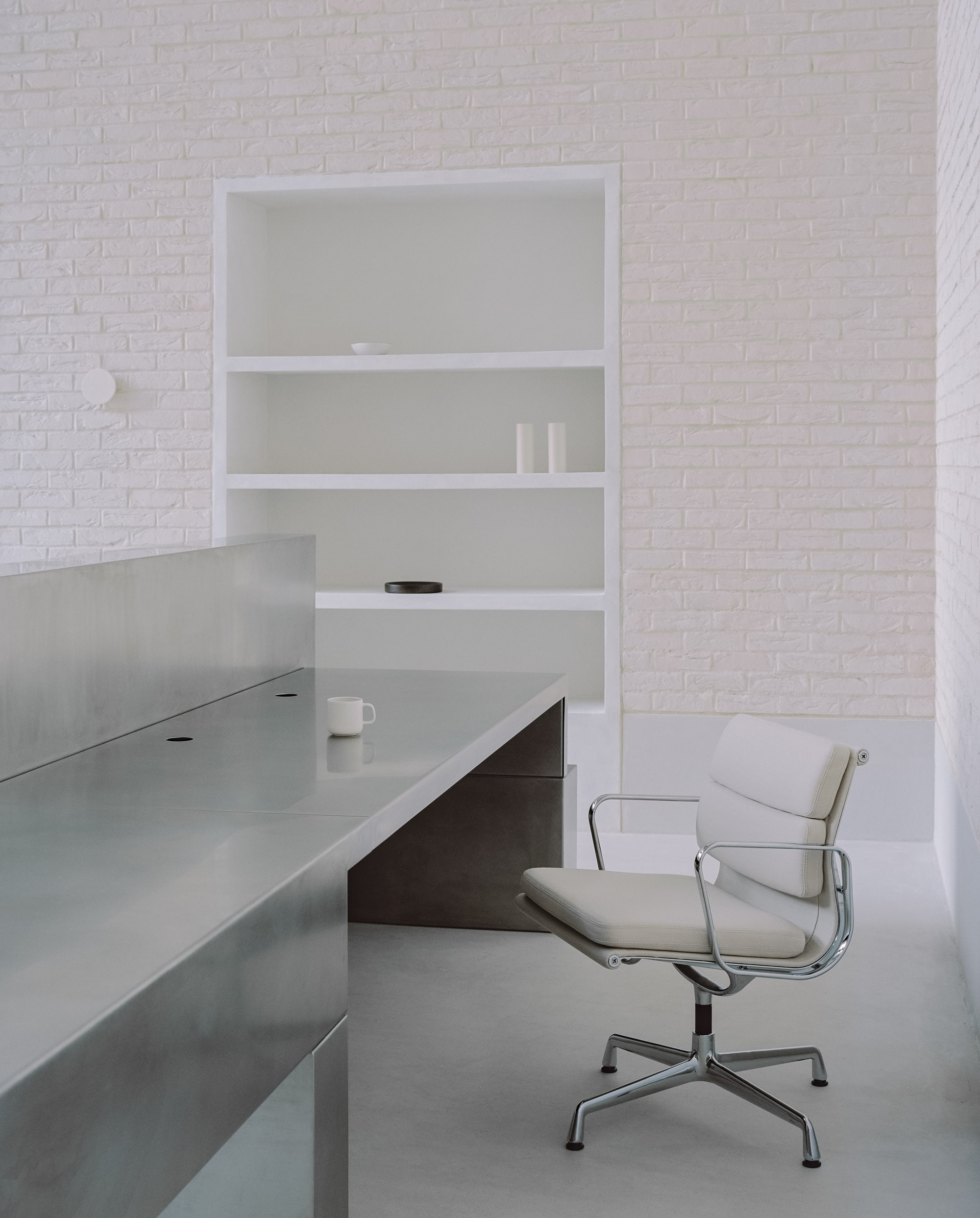 Close-up of a polished steel desk with recessed shelving and a leather office chair against white brick