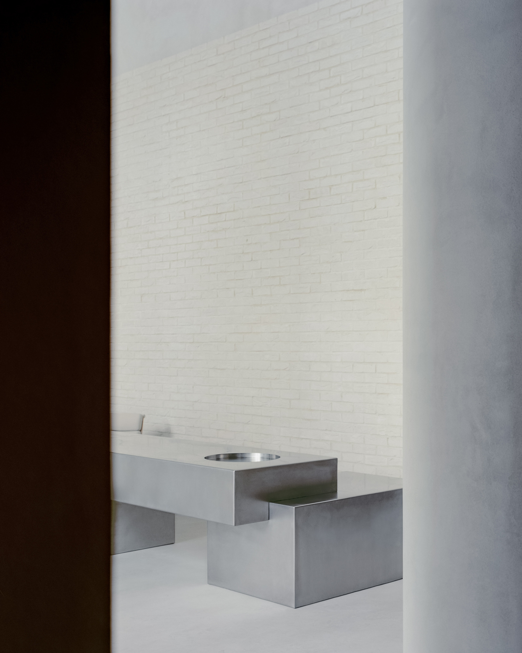 Stainless steel sink with circular basin inset into floating counter against white brick wall in shadow