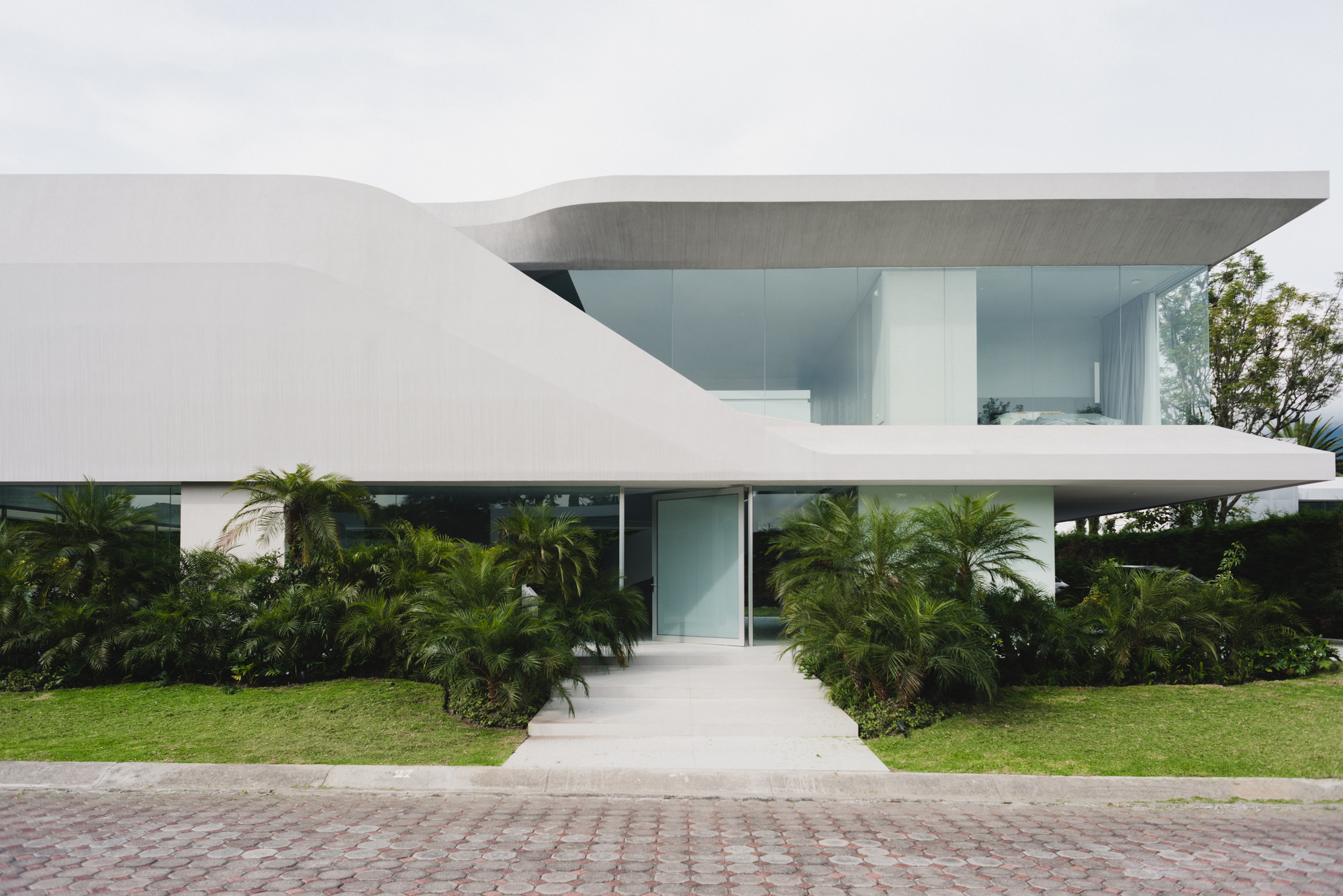 Street view of the white horizontal volumes with cantilevered upper floor and palm plantings in the lawn