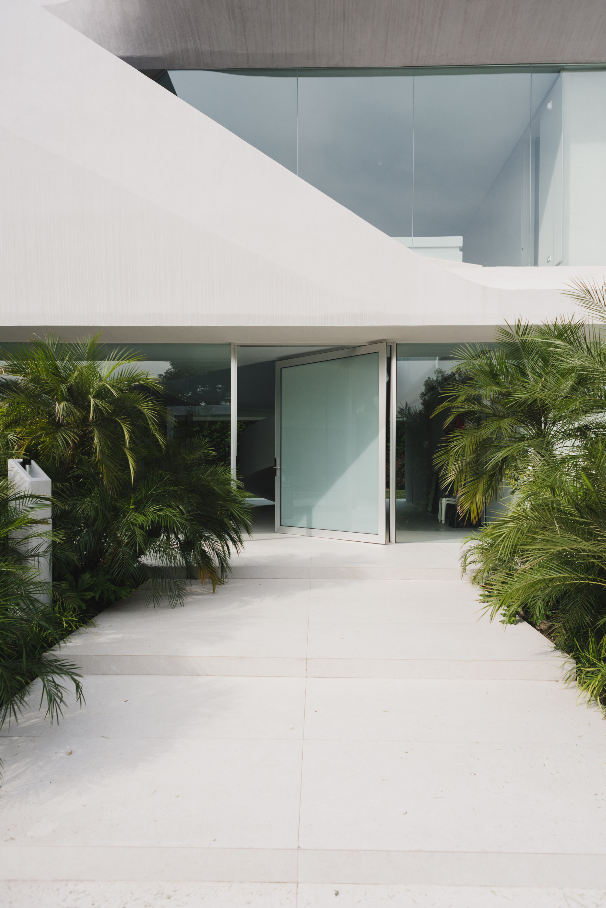 Entrance pathway lined with low palms leading to a pivoting glass door beneath white cantilevered slabs