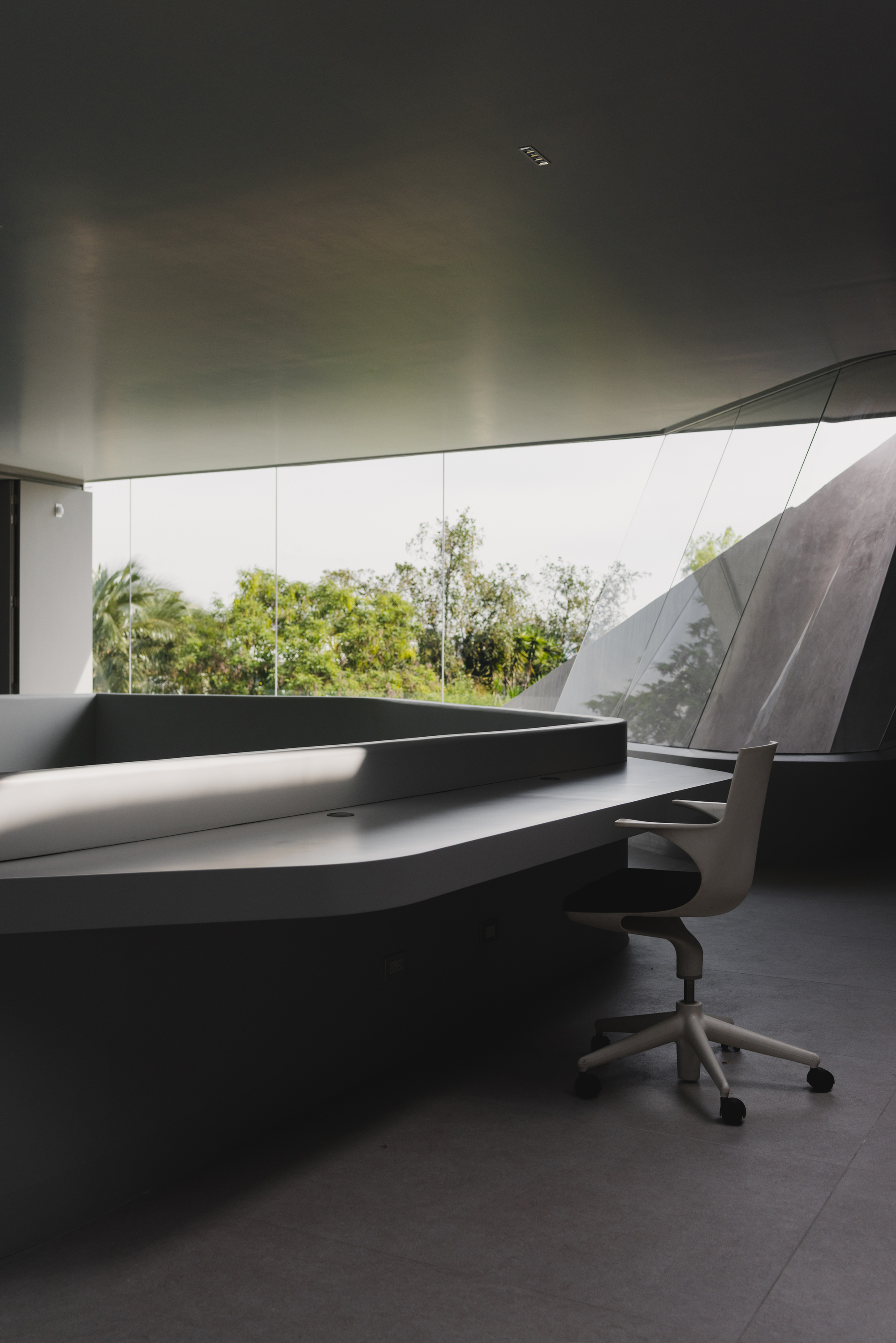 Dark desk workspace beneath a curved soffit with frameless glass opening to trees beyond