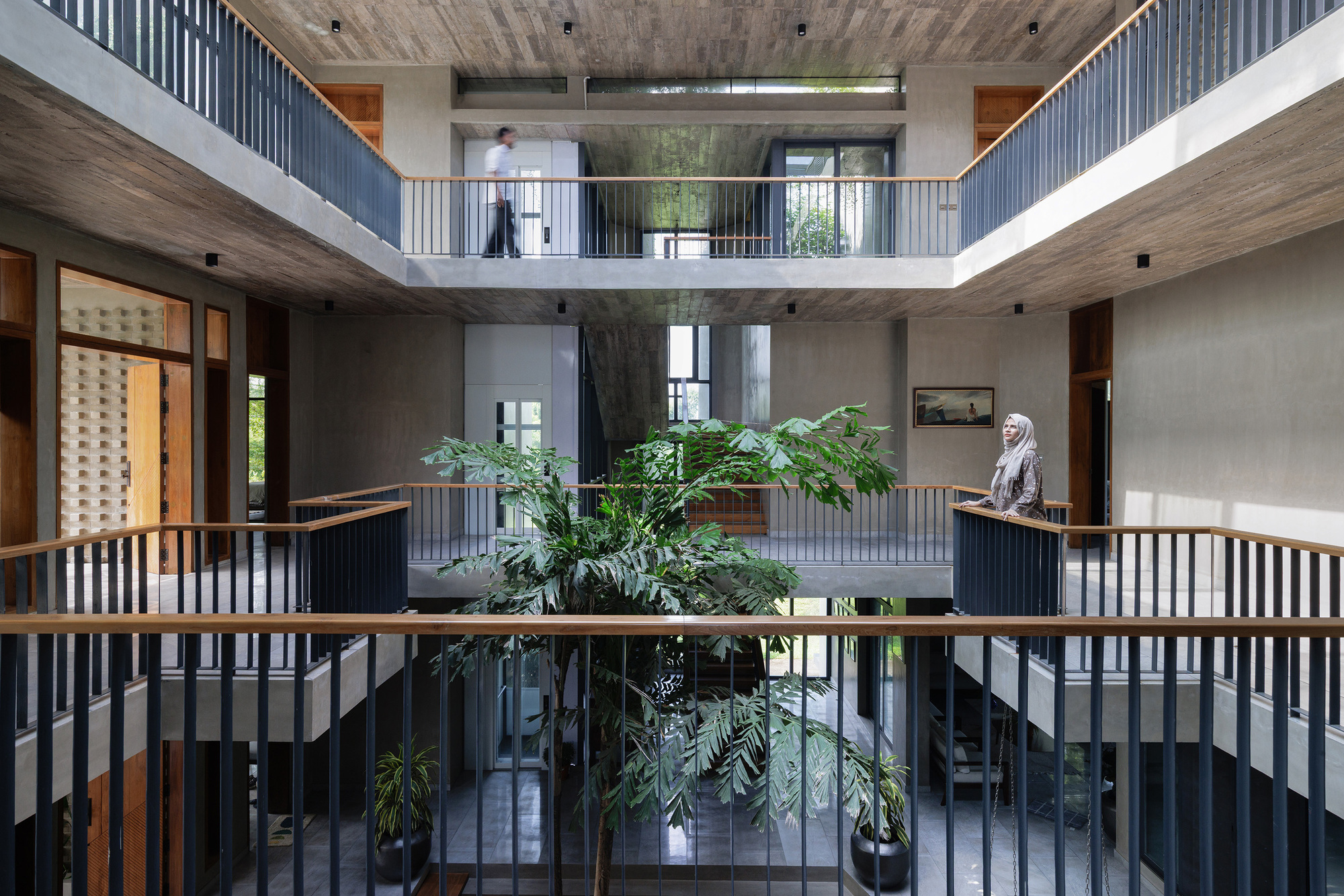 Three-story atrium with vertical slat balustrades and central tree planters with figure standing on upper gallery