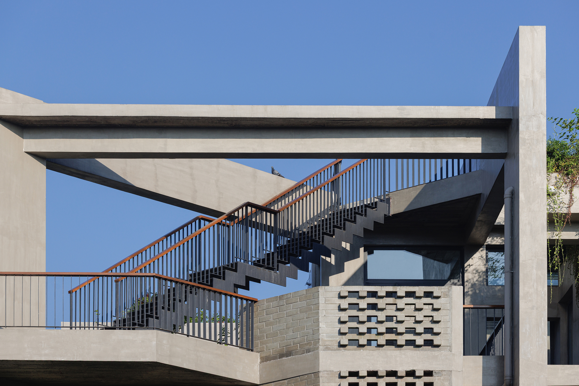 Exterior staircase with metal handrails ascending between concrete beams and perforated block screens under blue sky