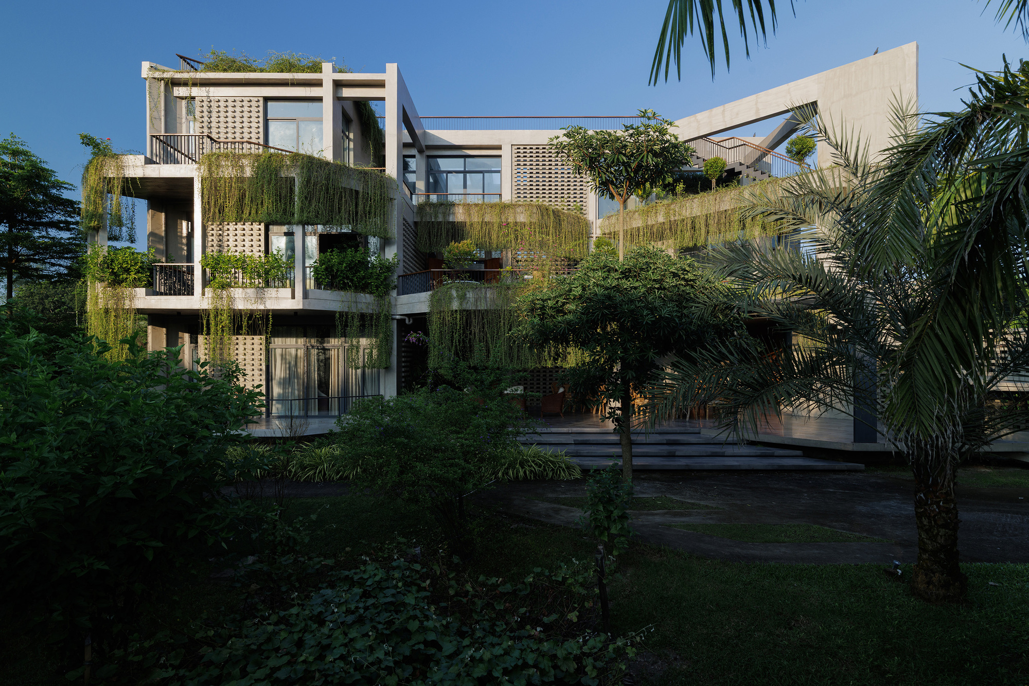 Garden facade showing stacked balconies with perforated concrete screens and hanging greenery among palm trees at dusk