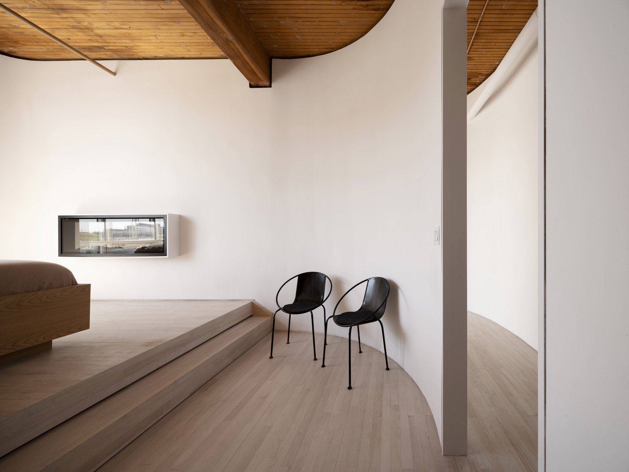 Curved platform with two black chairs beside a low horizontal window under vaulted timber ceiling