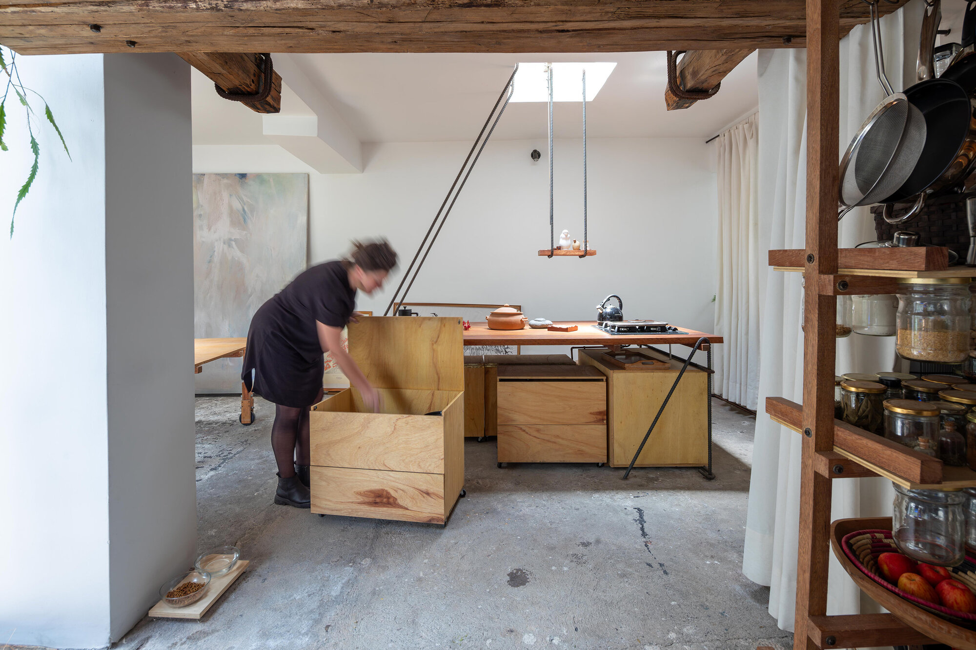 Open kitchen with plywood cabinetry under exposed timber beams and a person reaching into storage