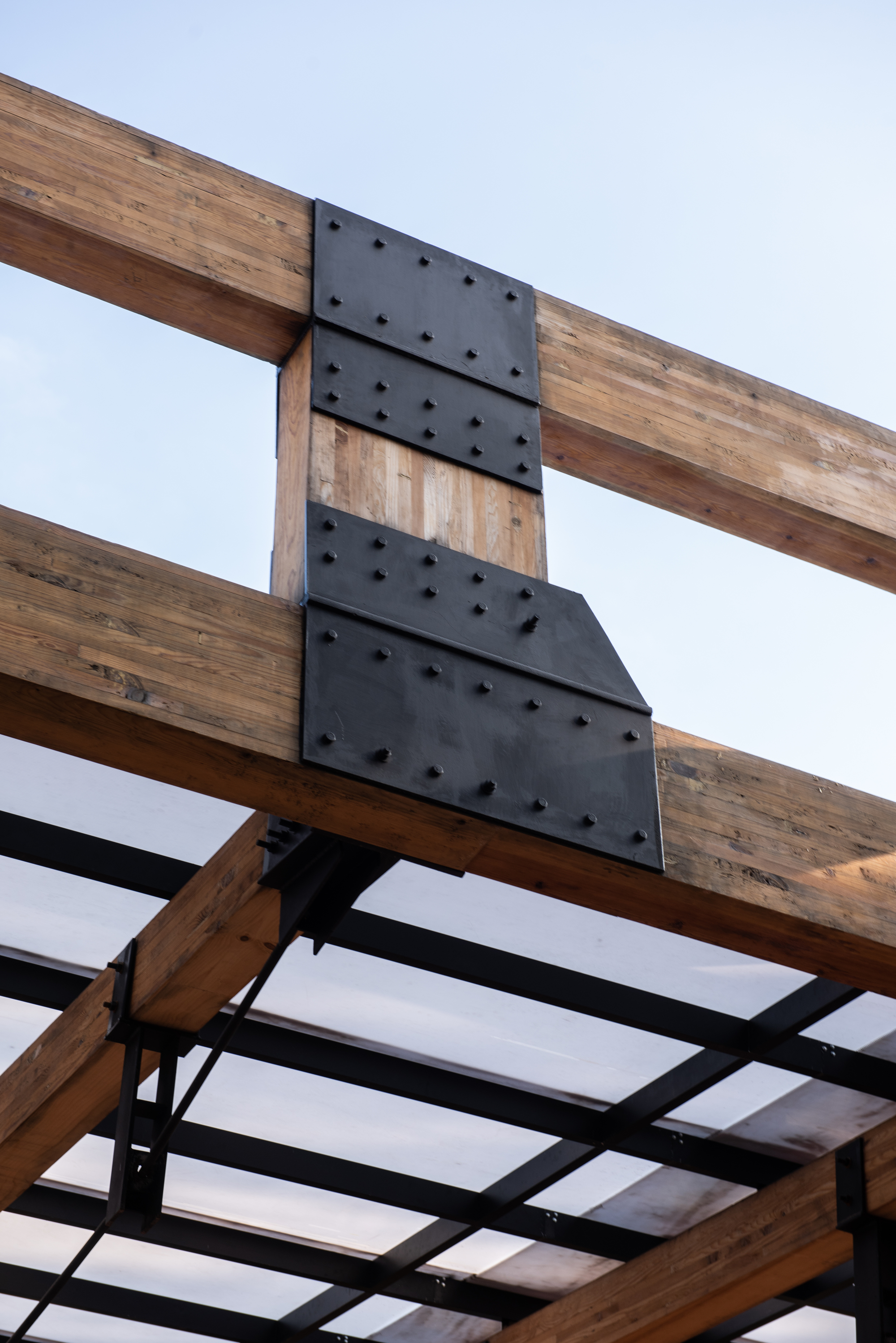 Close-up of a black bolted steel plate joint between laminated timber beams against a pale blue sky