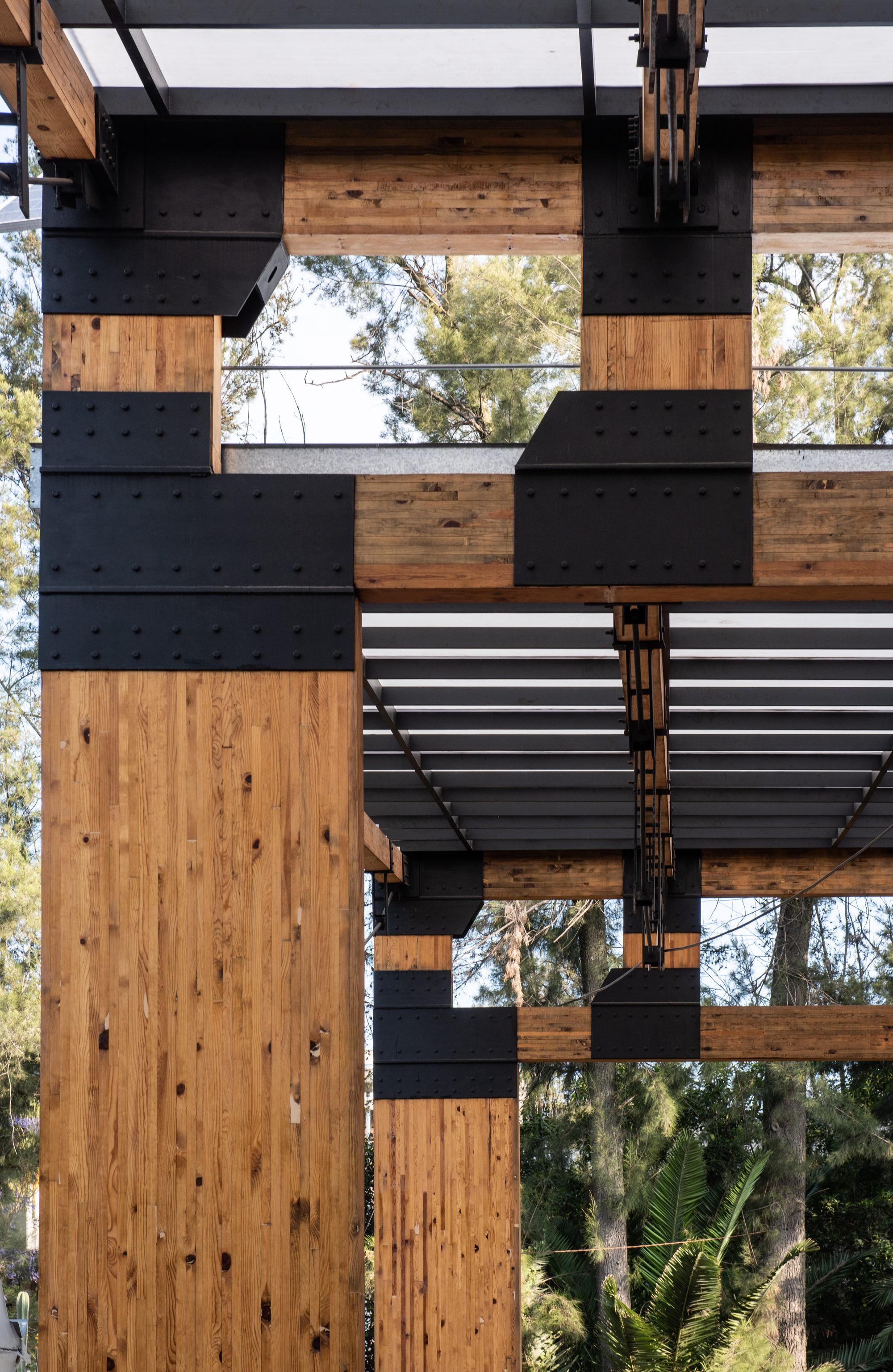 Close-up of riveted black steel plate connections joining laminated timber columns and beams, with pine trees behind