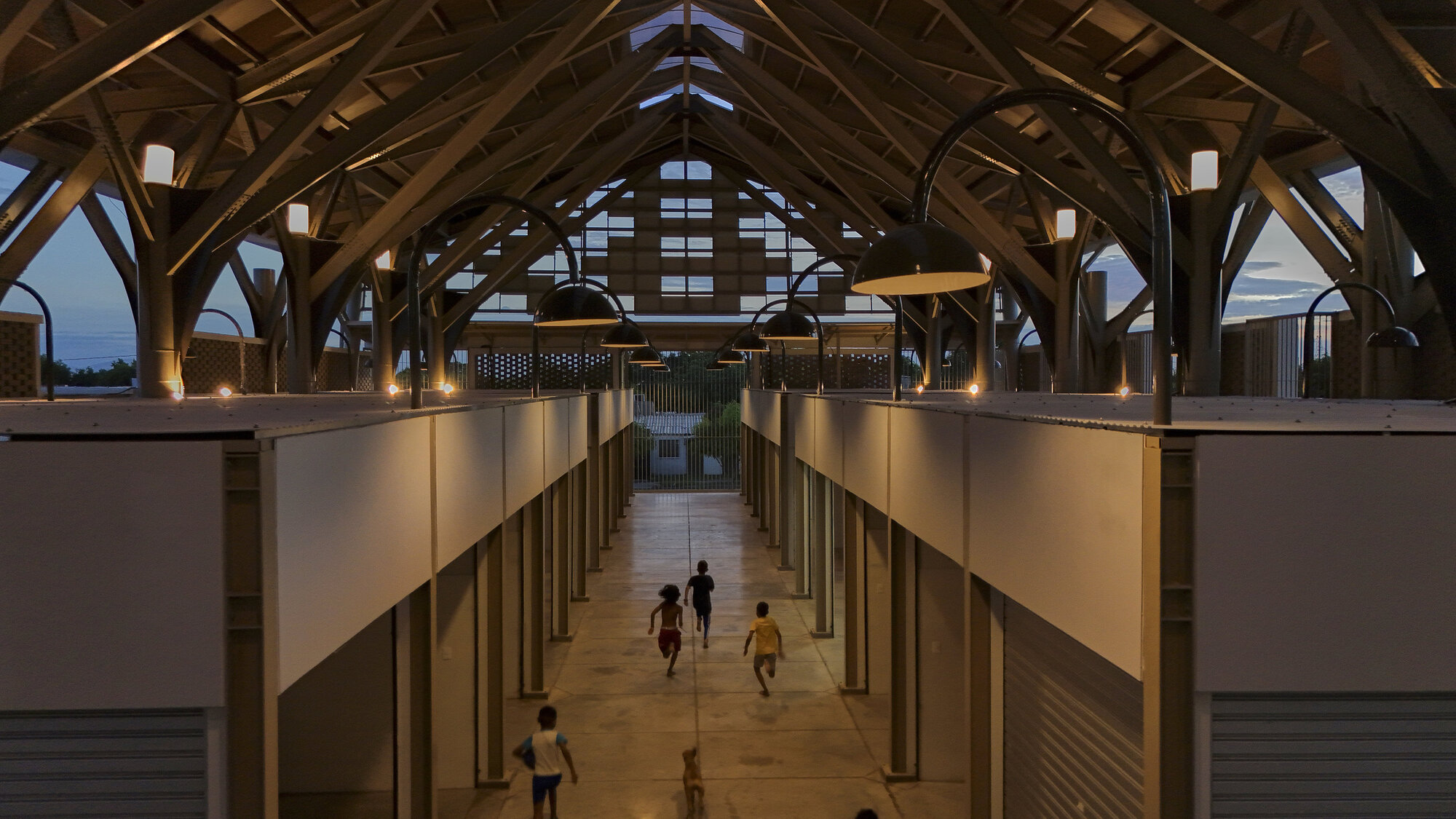 Interior corridor beneath an exposed timber frame roof with children walking between flanking volumes at dusk