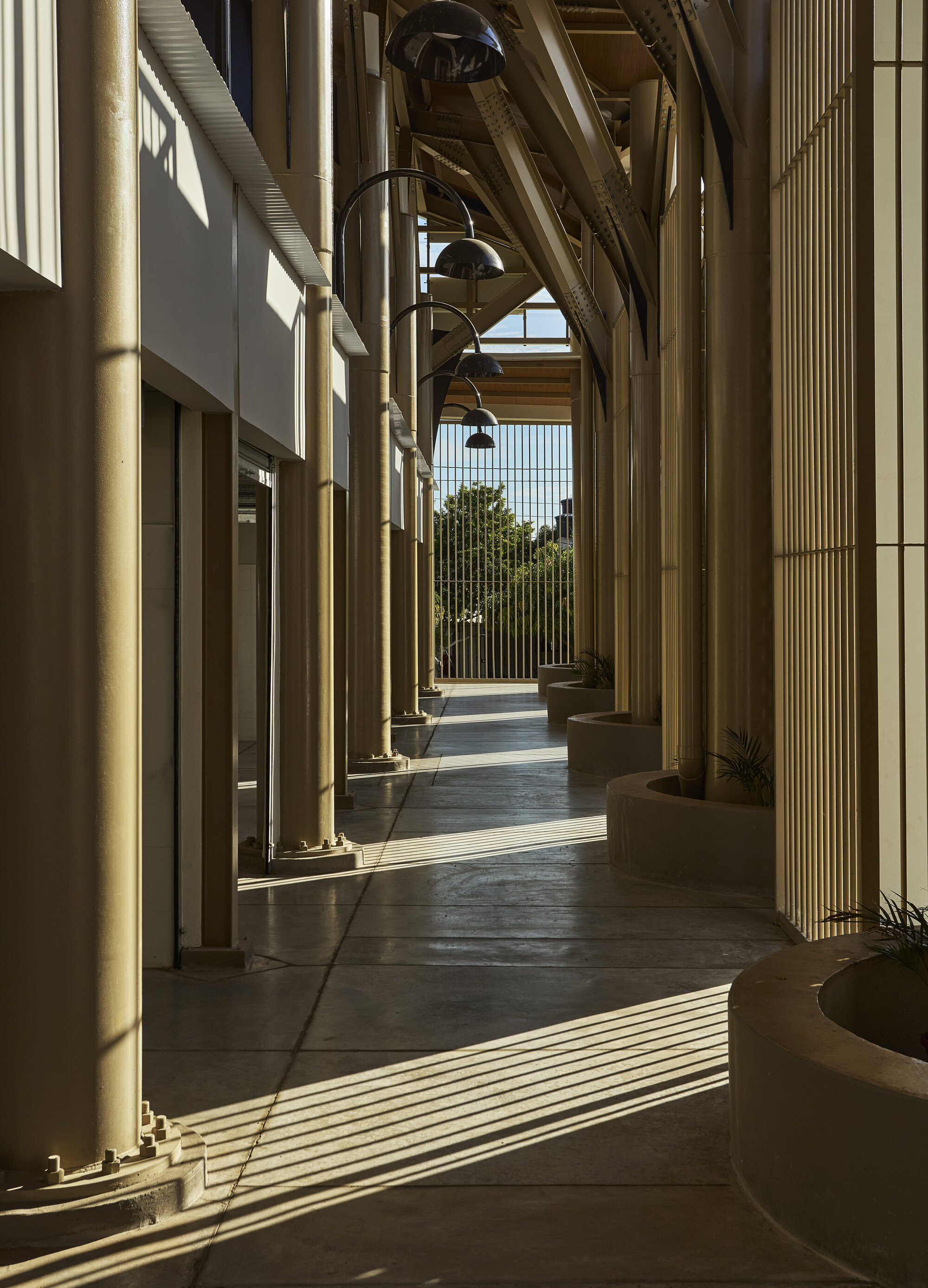 Covered walkway with slender cylindrical columns, pendant lights, and diagonal shadows striping the polished floor