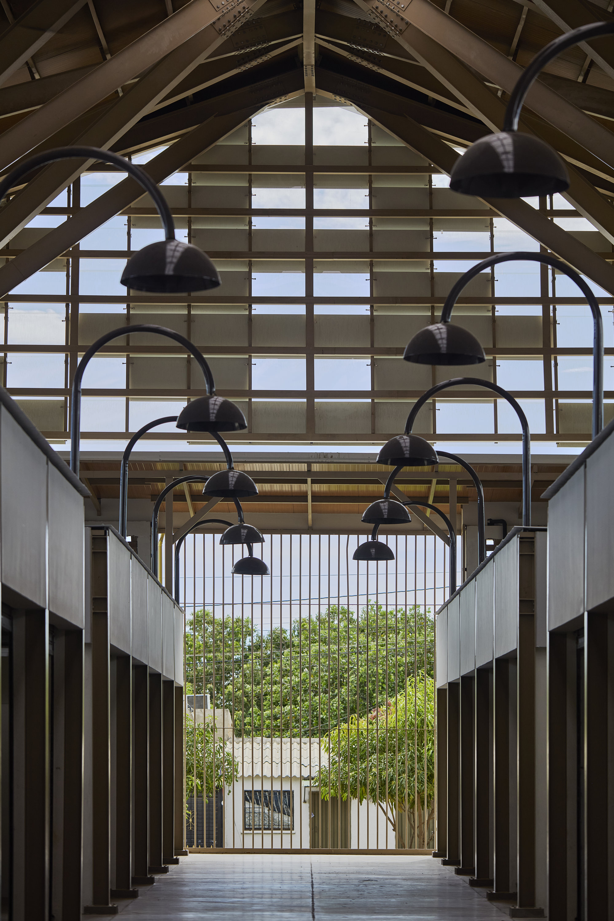 Interior arcade with exposed timber trusses, arched pendant lamps, and translucent end wall framing trees beyond