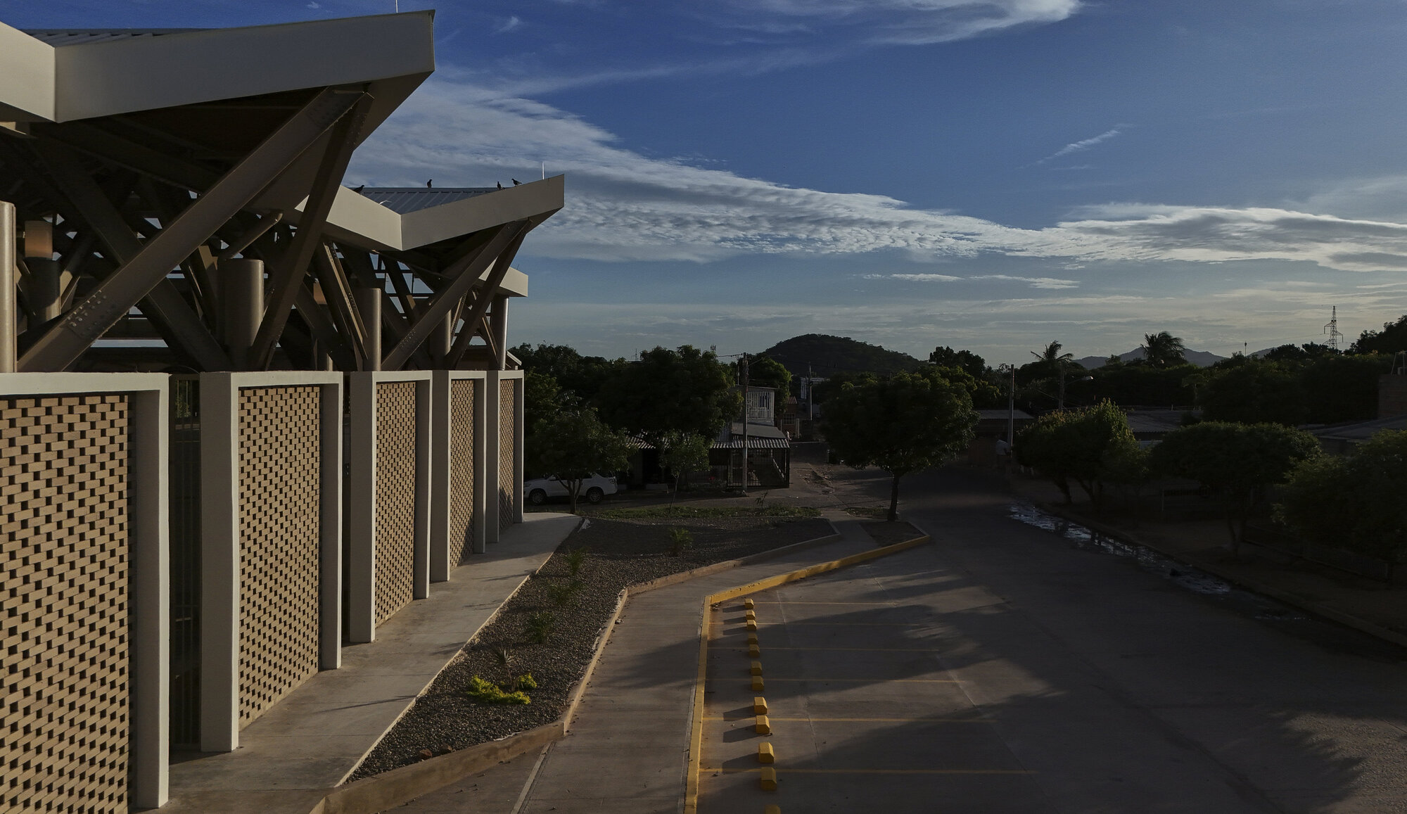 Covered walkway with timber-braced canopy, perforated screens, and view along the curved roadway at dusk
