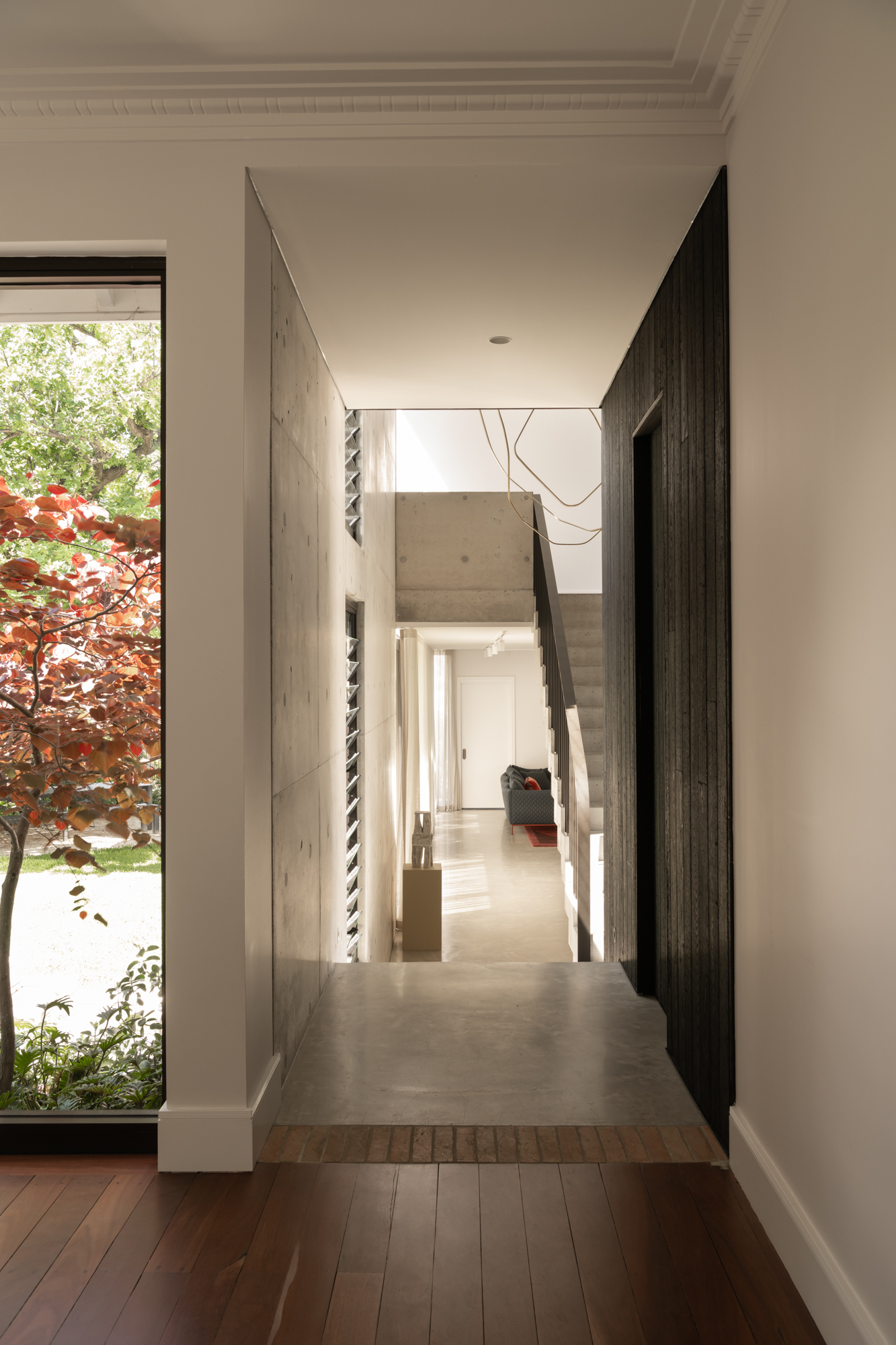 Hallway with concrete floor and charred timber walls looking through to a courtyard window framing red foliage