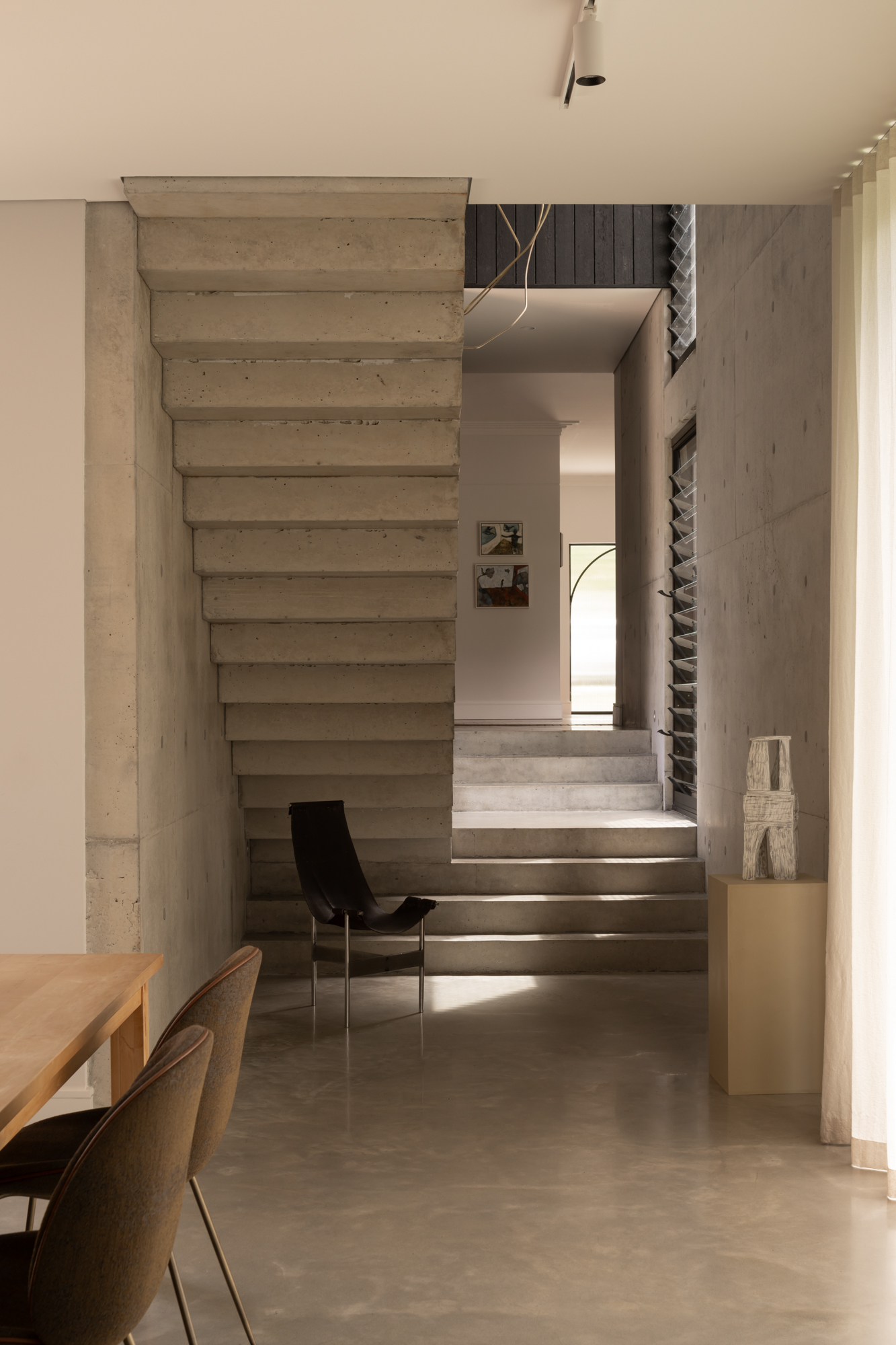 Board-formed concrete staircase with adjacent passage leading to a sunlit opening beyond a black chair