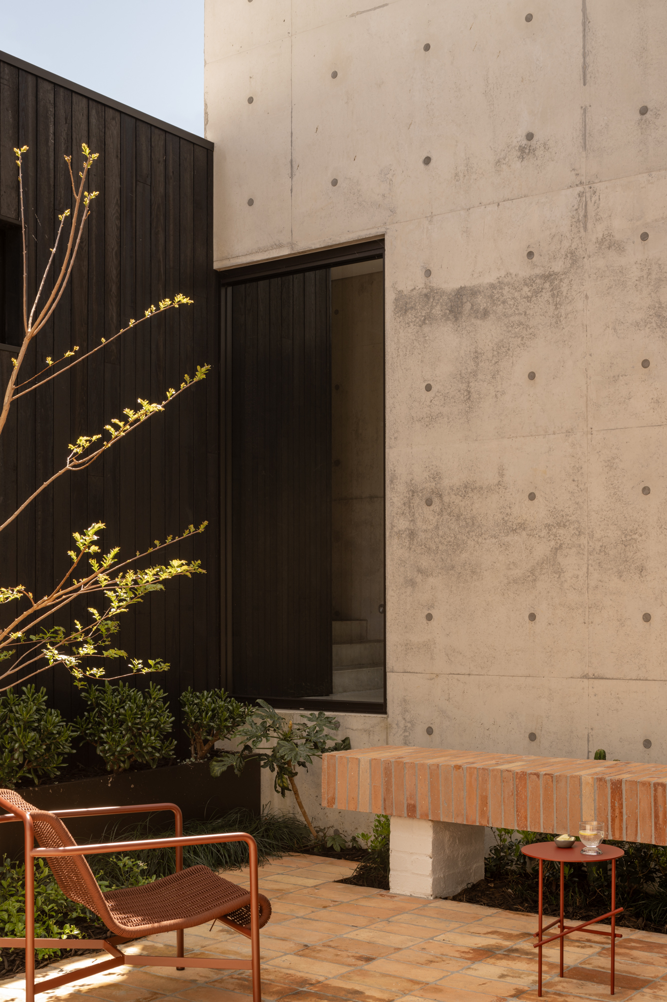 Board-formed concrete wall meeting timber cladding with doorway and terracotta brick bench