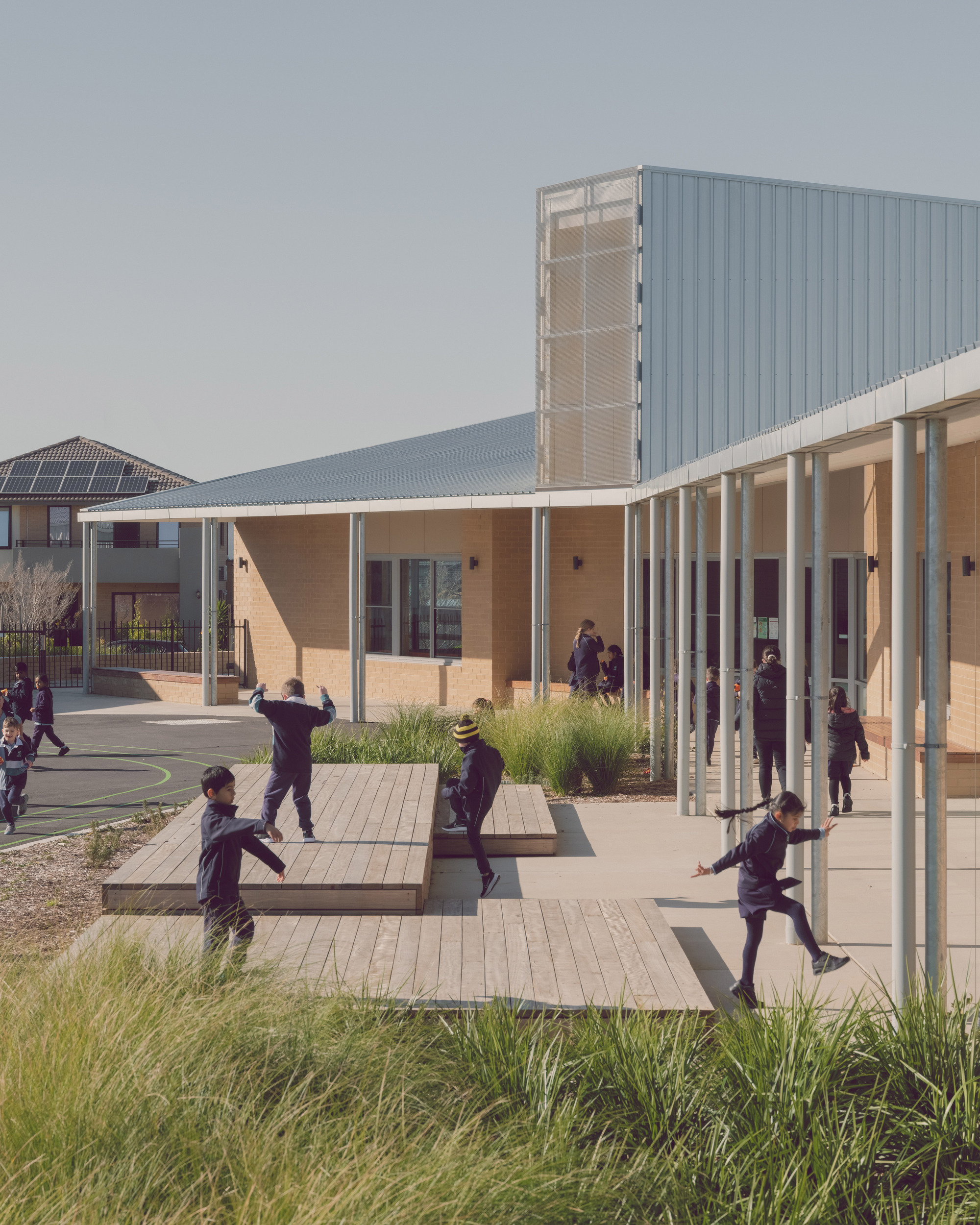 Covered walkway with slender white columns sheltering students playing on timber deck platforms and paving