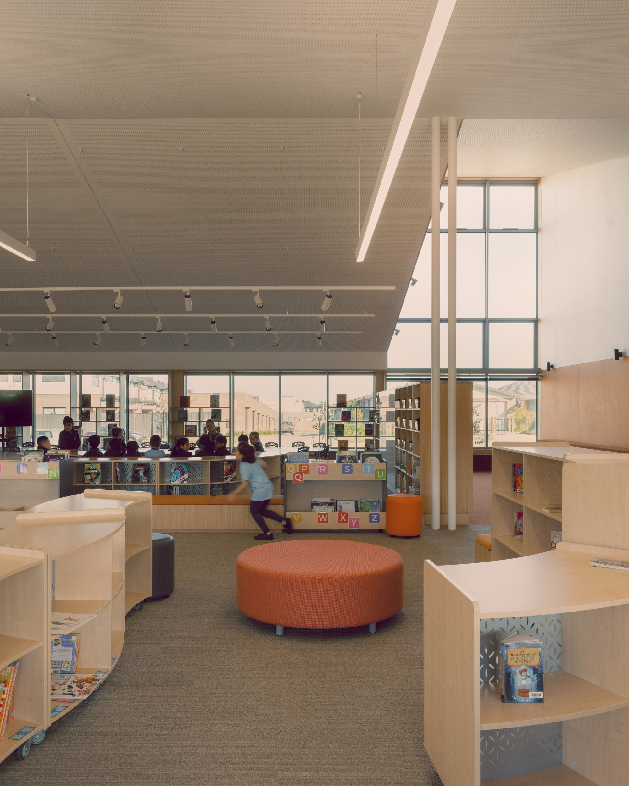 Double-height library interior with perforated ceiling panels, clerestory windows and students browsing low plywood shelving