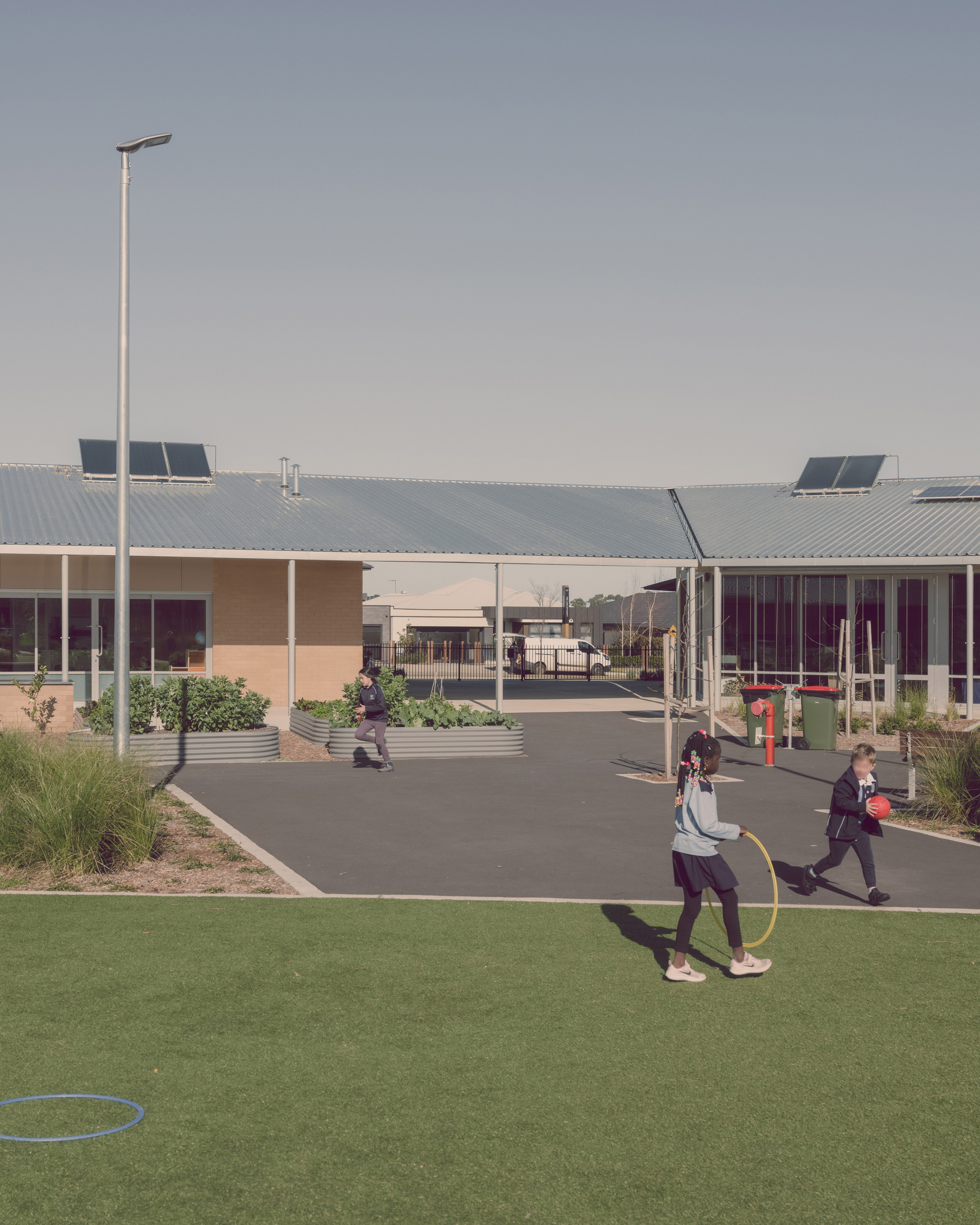 Courtyard view with children playing on synthetic turf and asphalt paths between low-slung buildings with corrugated metal roofs and solar panels