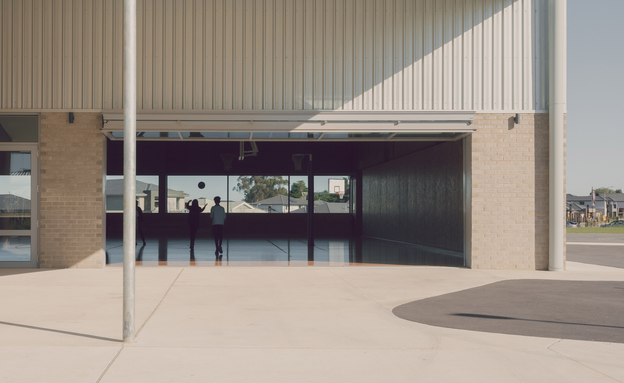 Covered breezeway with cream brick walls, vertical metal cladding and deep eaves framing views to the interior basketball court