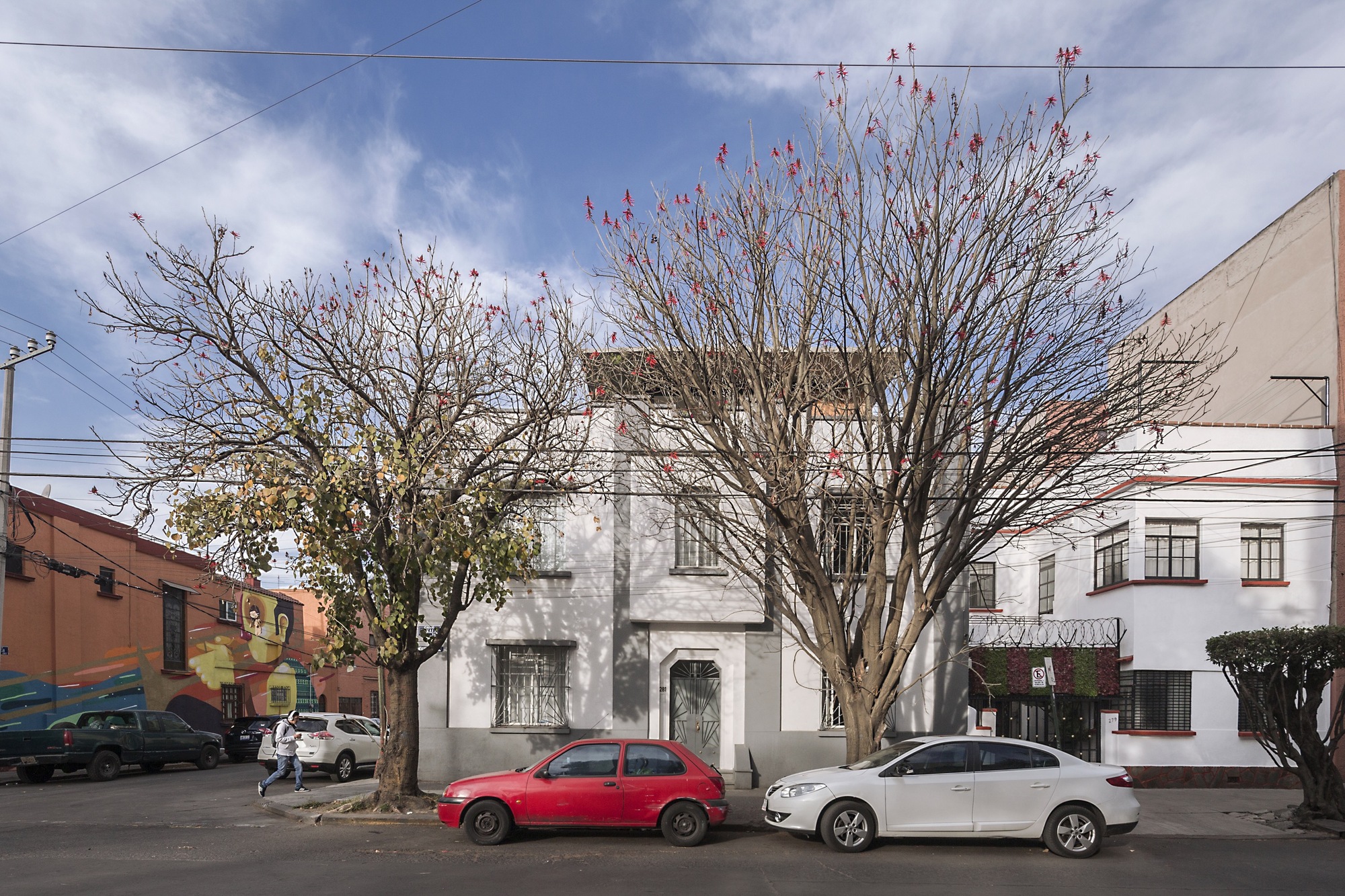 White street facade of a two-storey house behind two flowering bare trees, with a red car parked out front and a pedestrian passing under blue sky