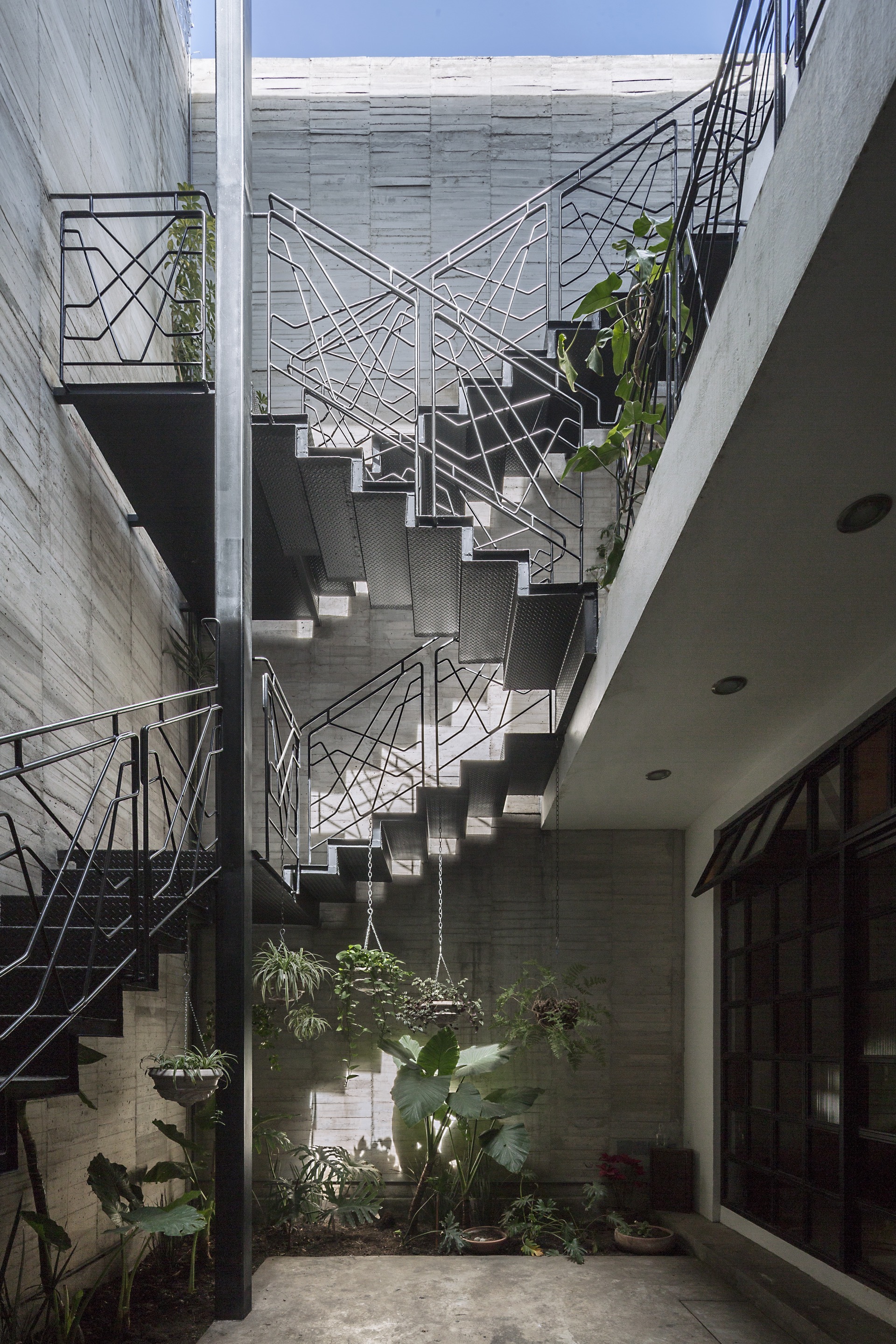 Steel staircase with geometric black railings zigzagging up a board-marked concrete light well, with potted ferns and hanging planters below
