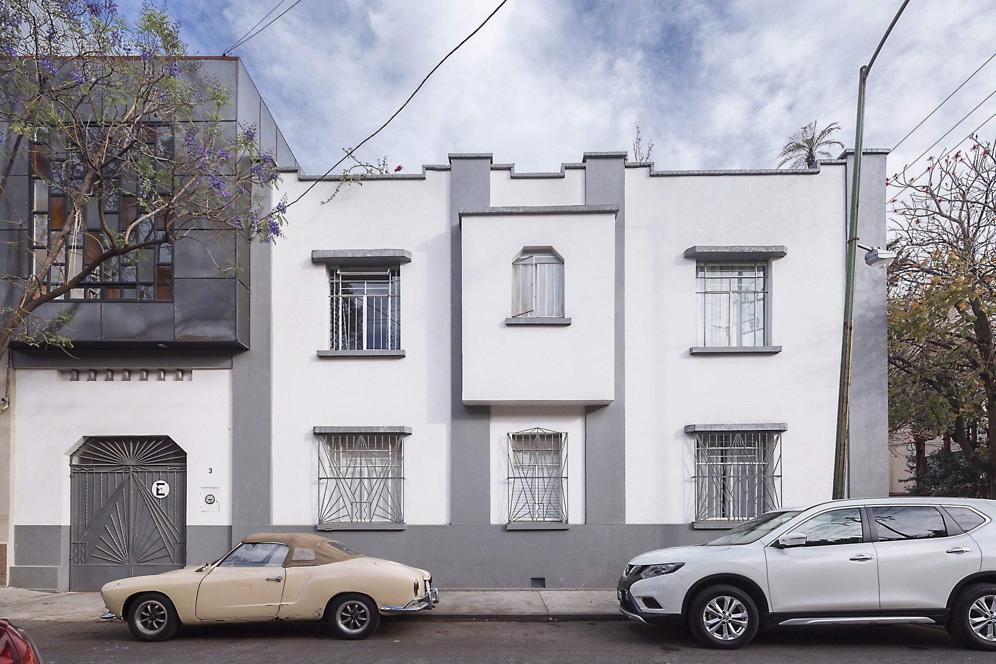 White and grey street facade with barred windows and a patterned metal garage door, a vintage cream convertible parked in front under cloudy sky