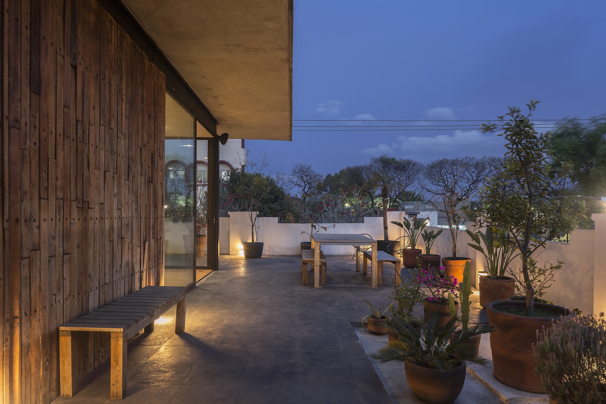 Dusk terrace with a reclaimed timber wall, a wooden bench, a long dining table and terracotta pots of plants along a white parapet