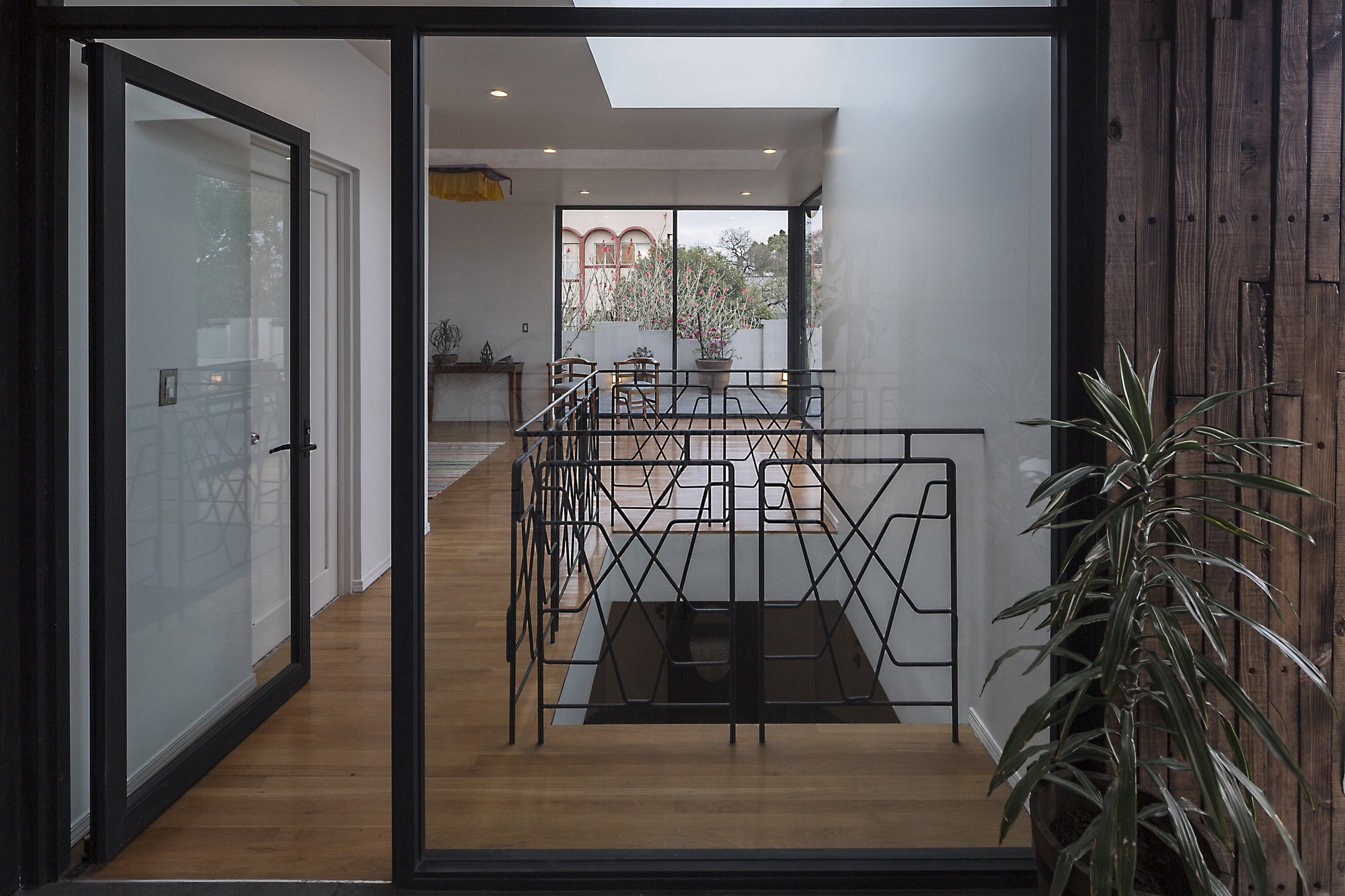 View through a black-framed glass door onto an upper hallway with wood floor, a geometric steel railing around a stairwell opening and a potted plant